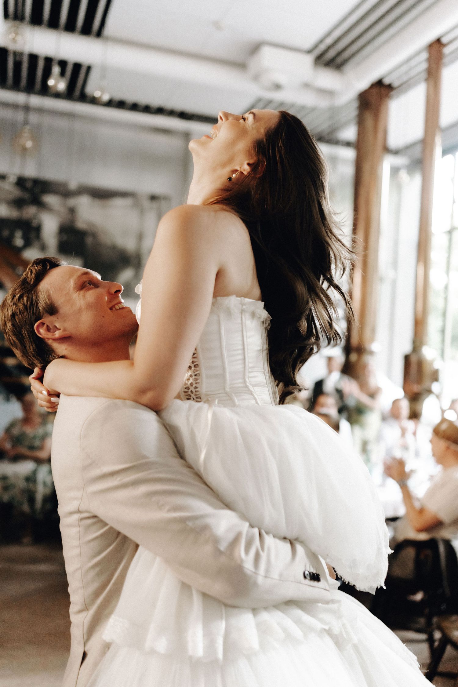 Groom lifts joyful bride in white tiered wedding gown during energetic first dance at reception.