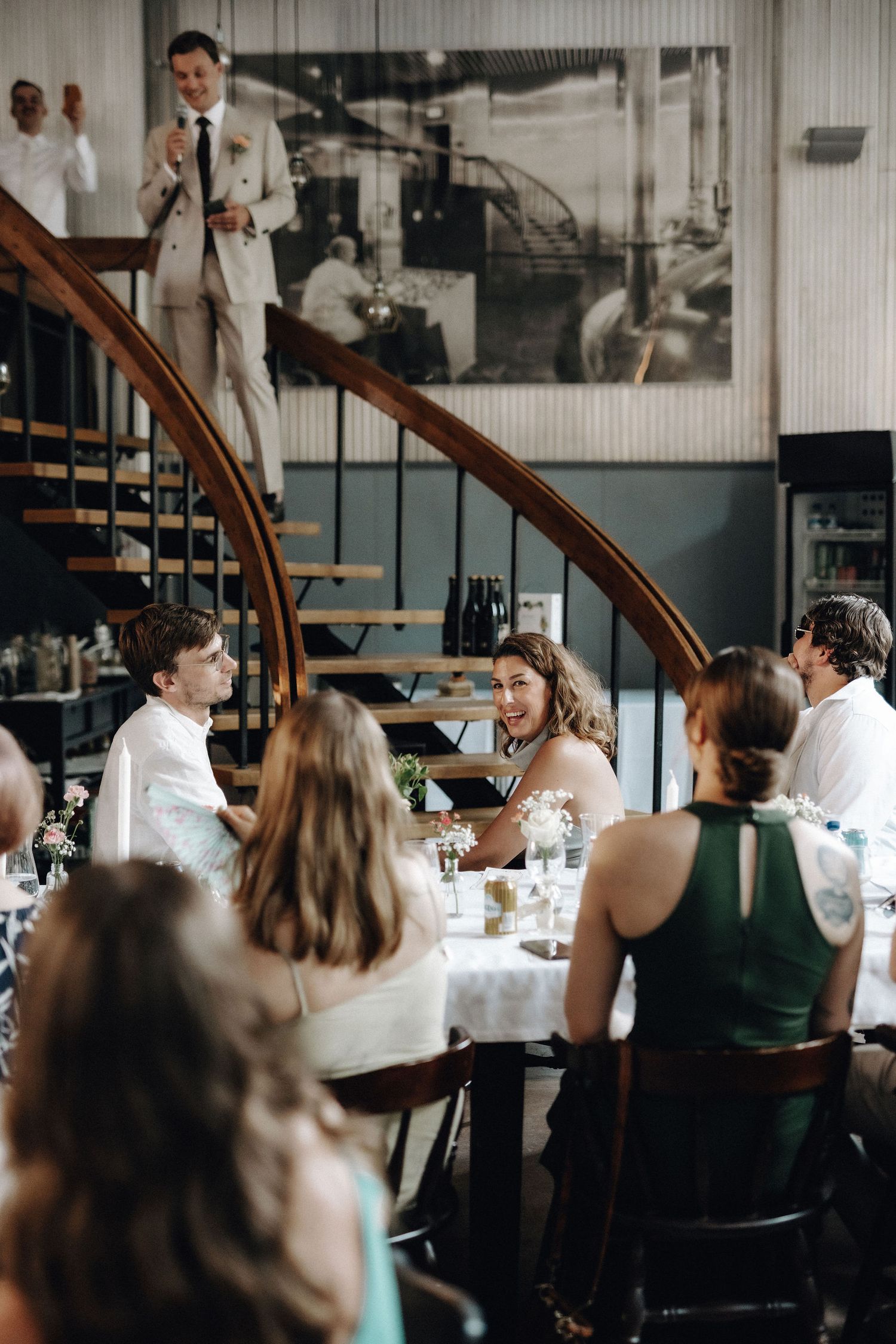 Bride laughing at table while groom delivers speech from spiral staircase above guests.