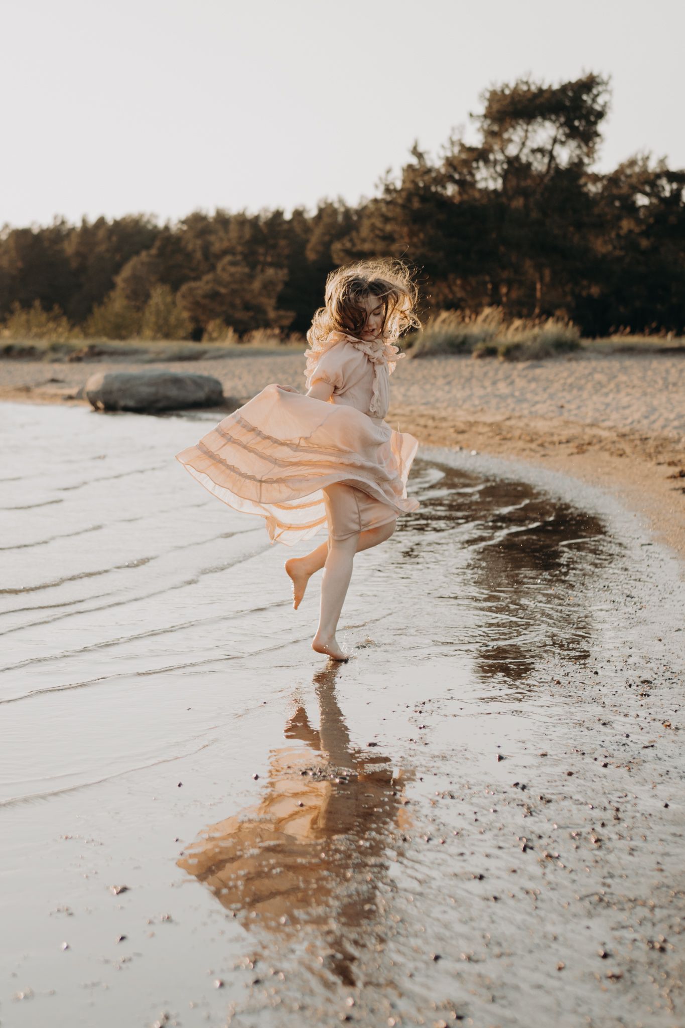A young girl in a flowing blush dress twirls and skips barefoot along a sandy beach at sunset.