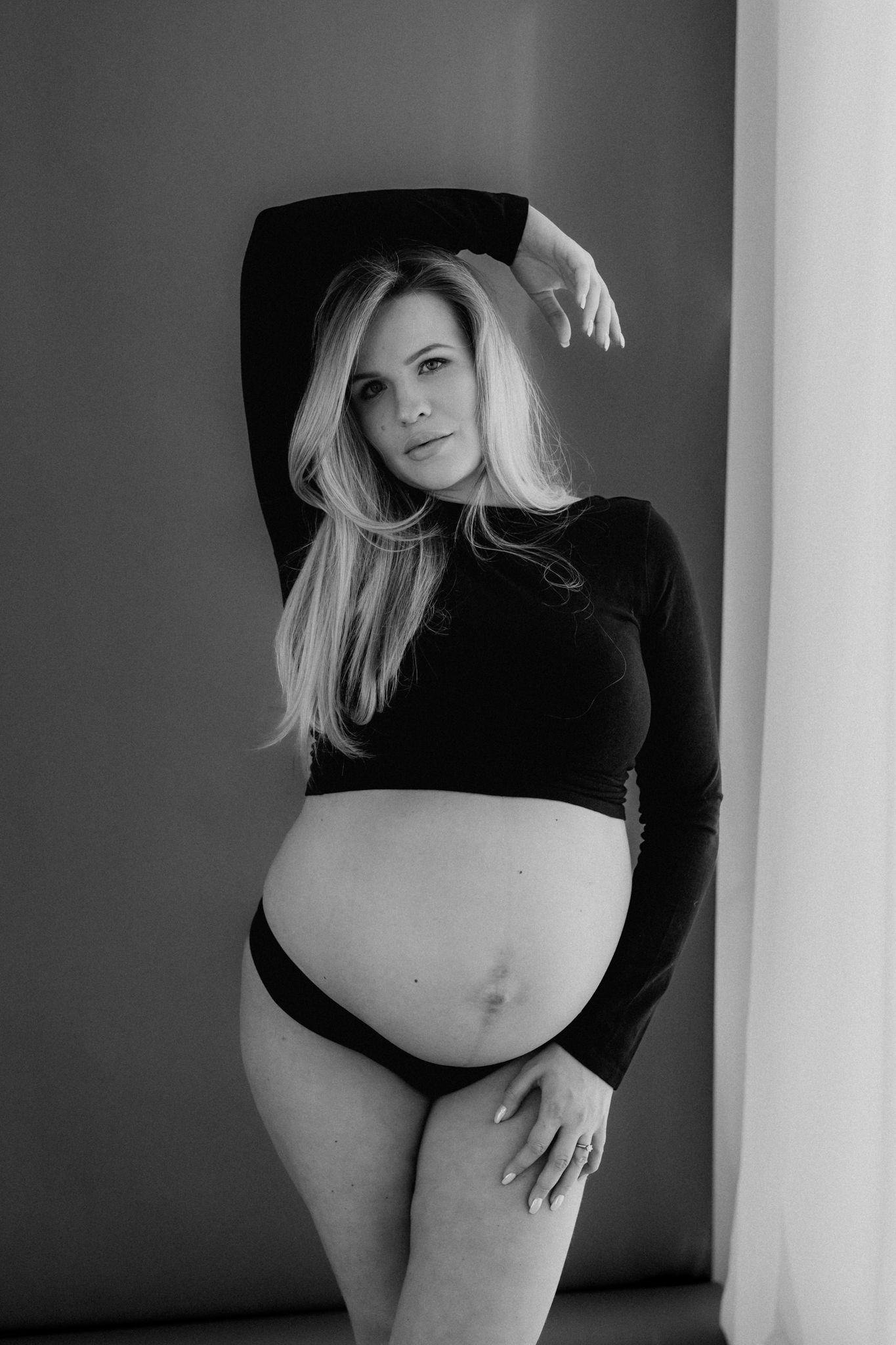 Black and white studio maternity photo of pregnant blonde woman in black crop top posing confidently.