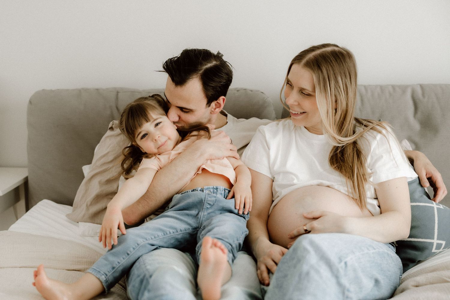 Father kissing young child while pregnant mother smiles sitting together on couch.