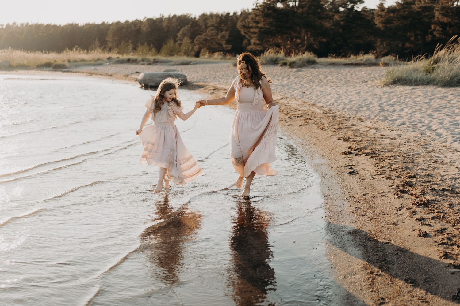 A mother and daughter in matching blush dresses hold hands while walking through gentle beach waves.