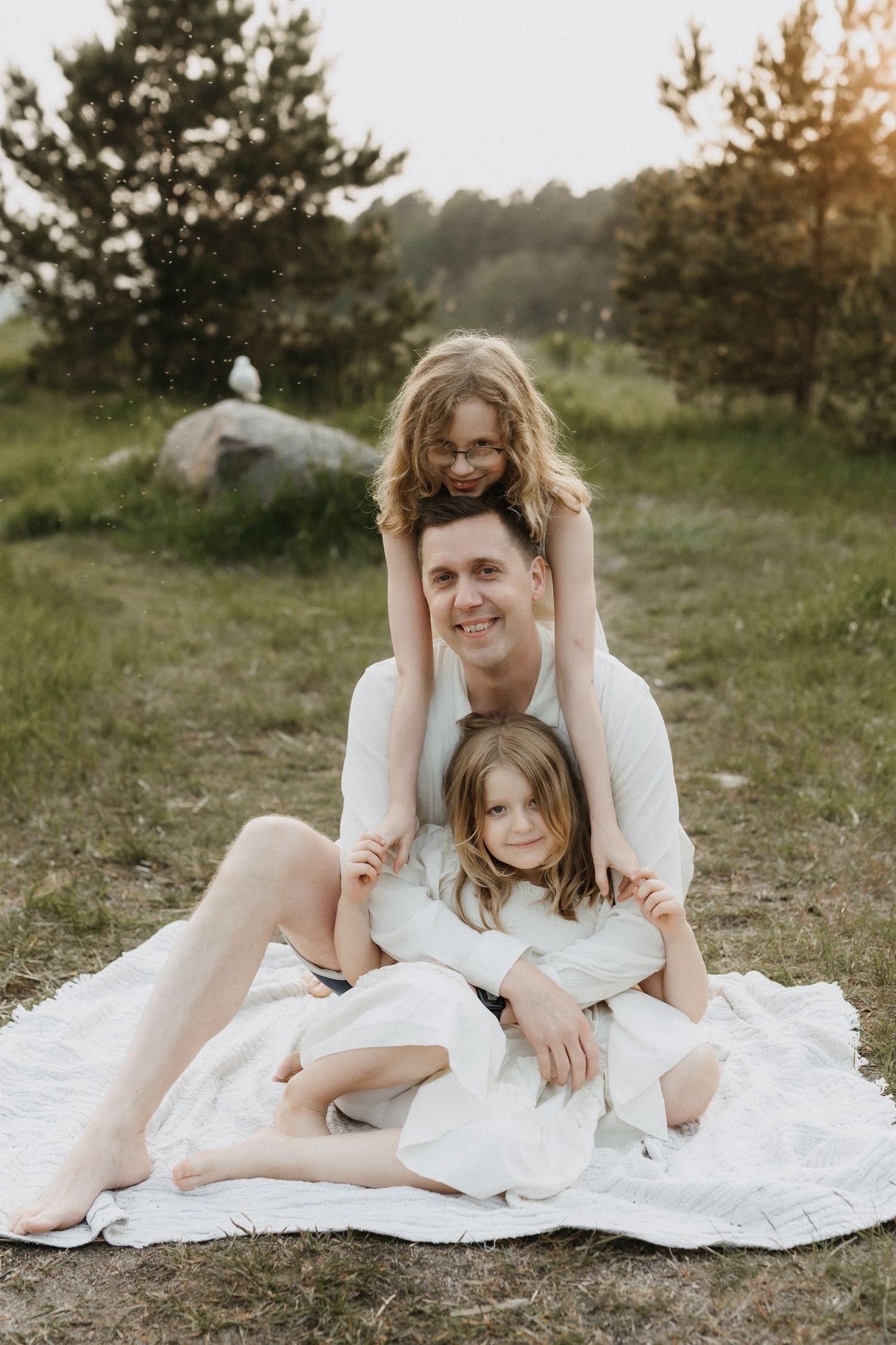 A smiling father sits on a blanket outdoors with his two daughters climbing on him playfully.