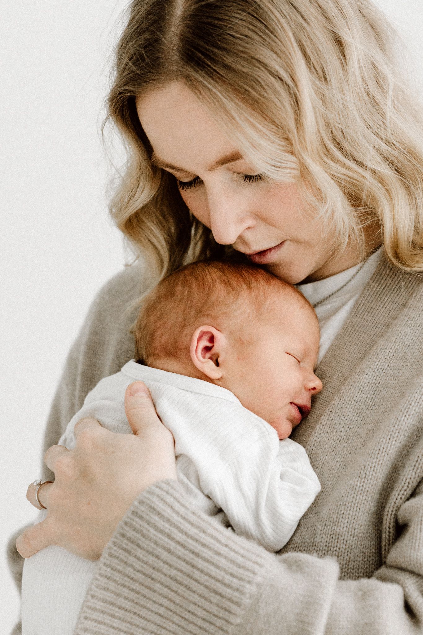 Blonde mother in beige knit cardigan gazing lovingly at her sleeping newborn baby.