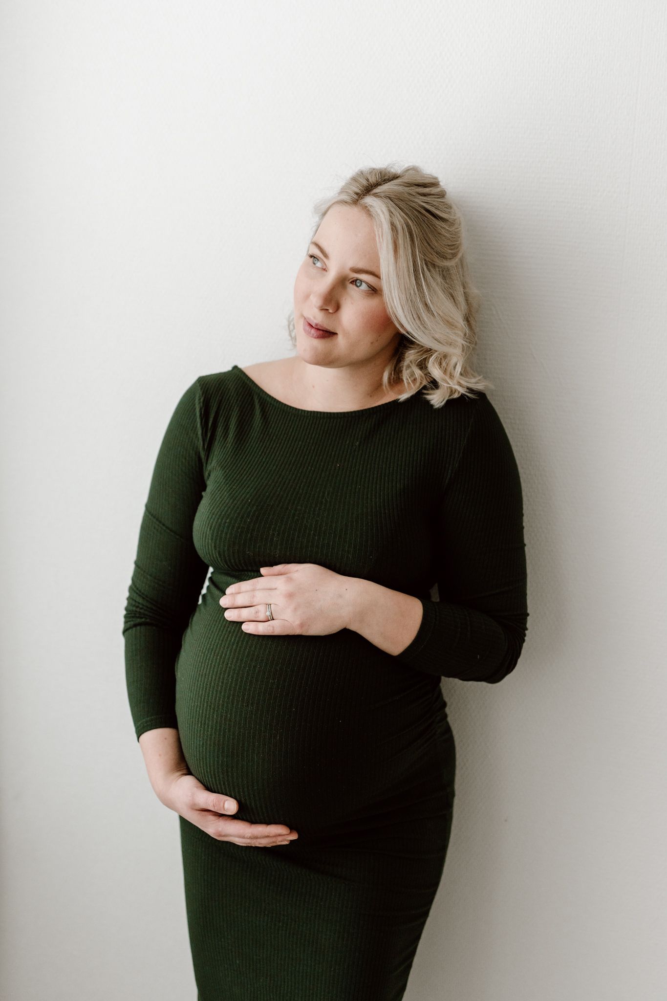 Blonde pregnant woman in forest green fitted dress holding belly while leaning against white wall.