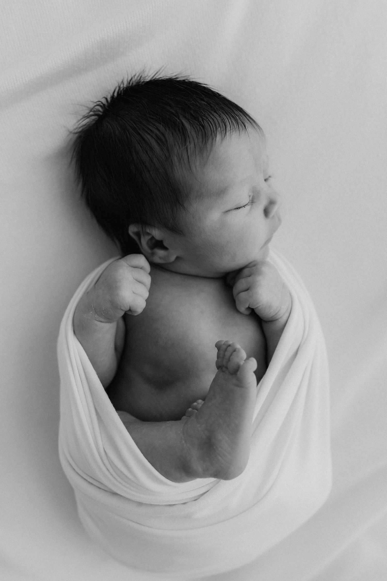 Black and white portrait of sleeping newborn baby wrapped in white swaddle blanket.