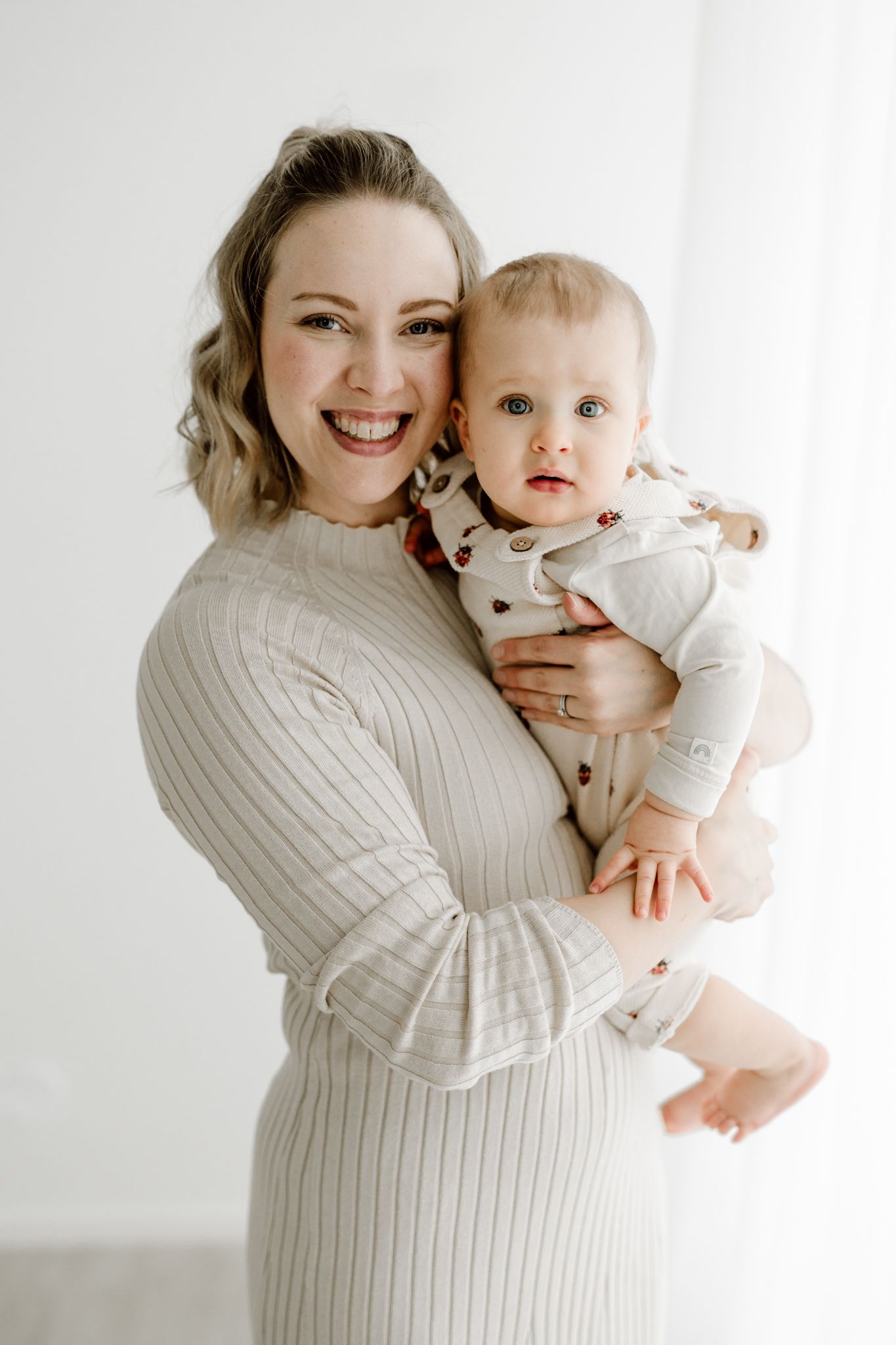 Smiling mother in a ribbed dress holding her wide-eyed baby against a white background.
