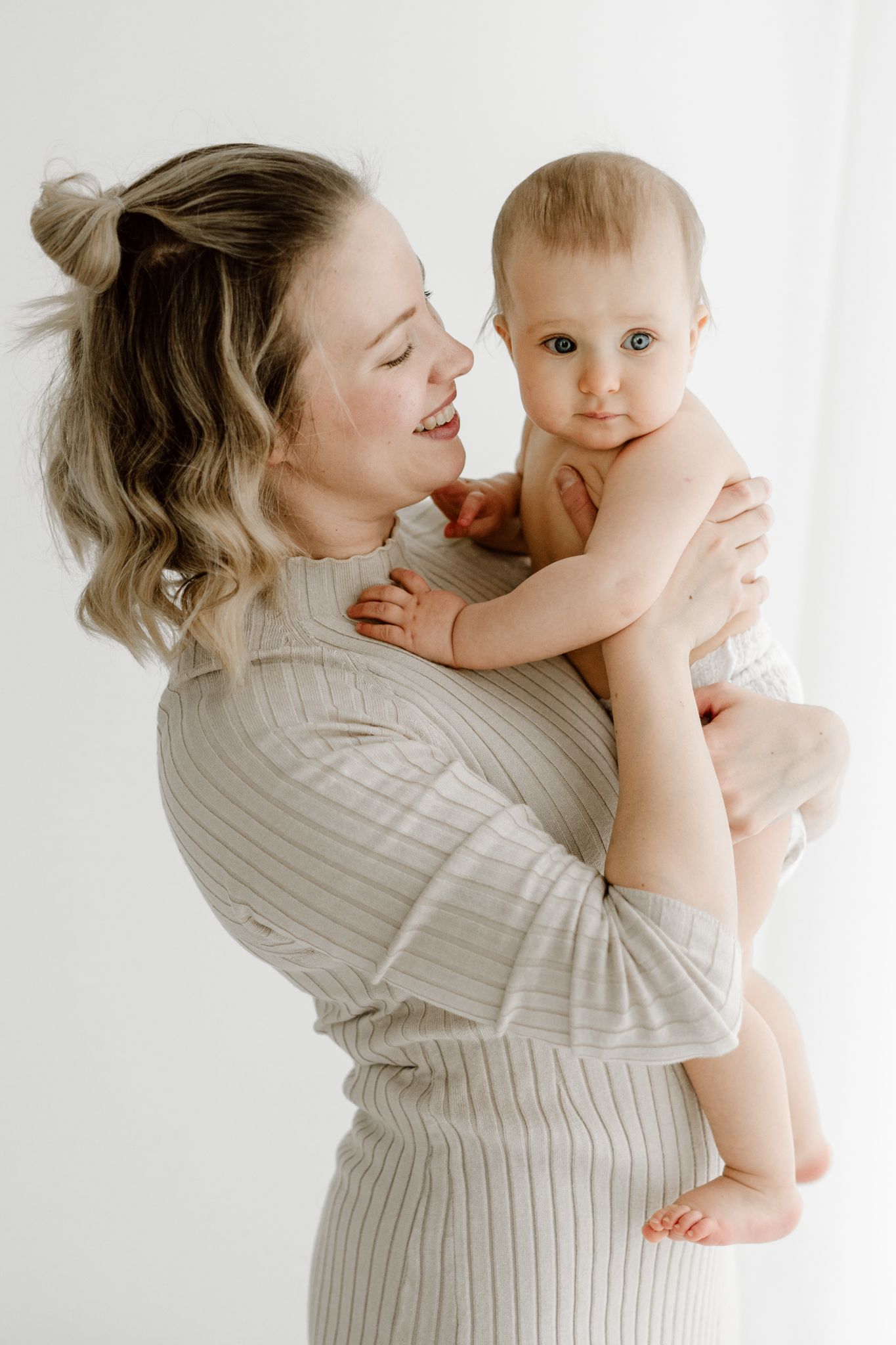 Happy mother in striped dress holding and gazing at her baby against a white background.