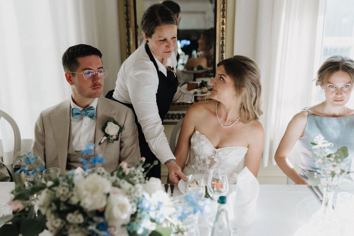 Bride in lace gown and groom in beige suit interact with guest at decorated wedding table.
