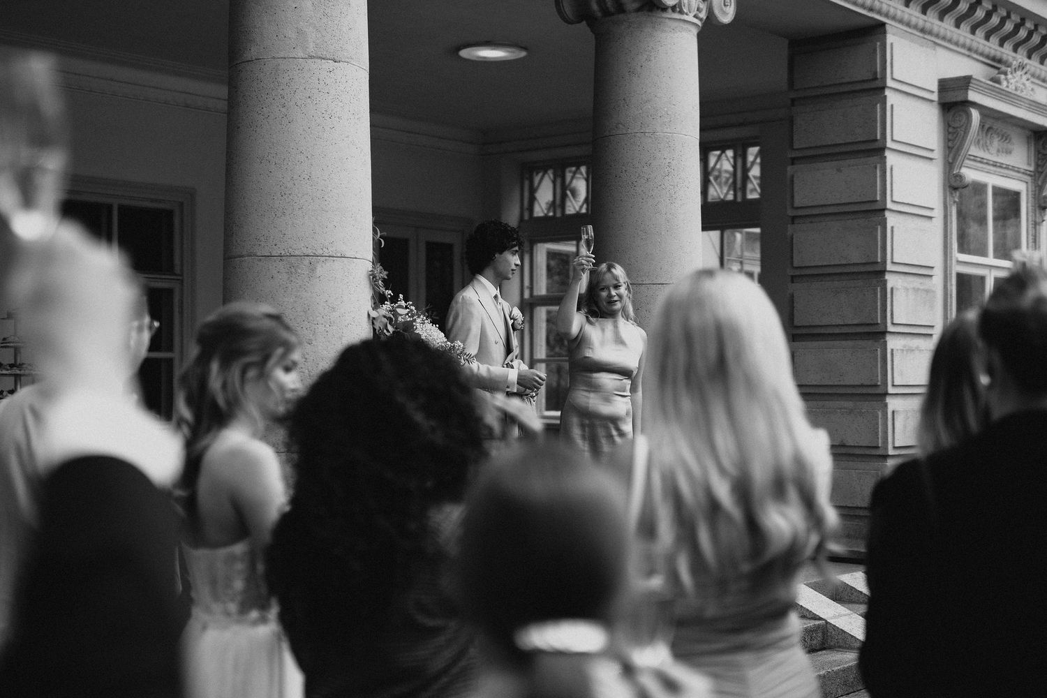 Black and white photo of wedding guests watching a couple exchange vows at a columned building.