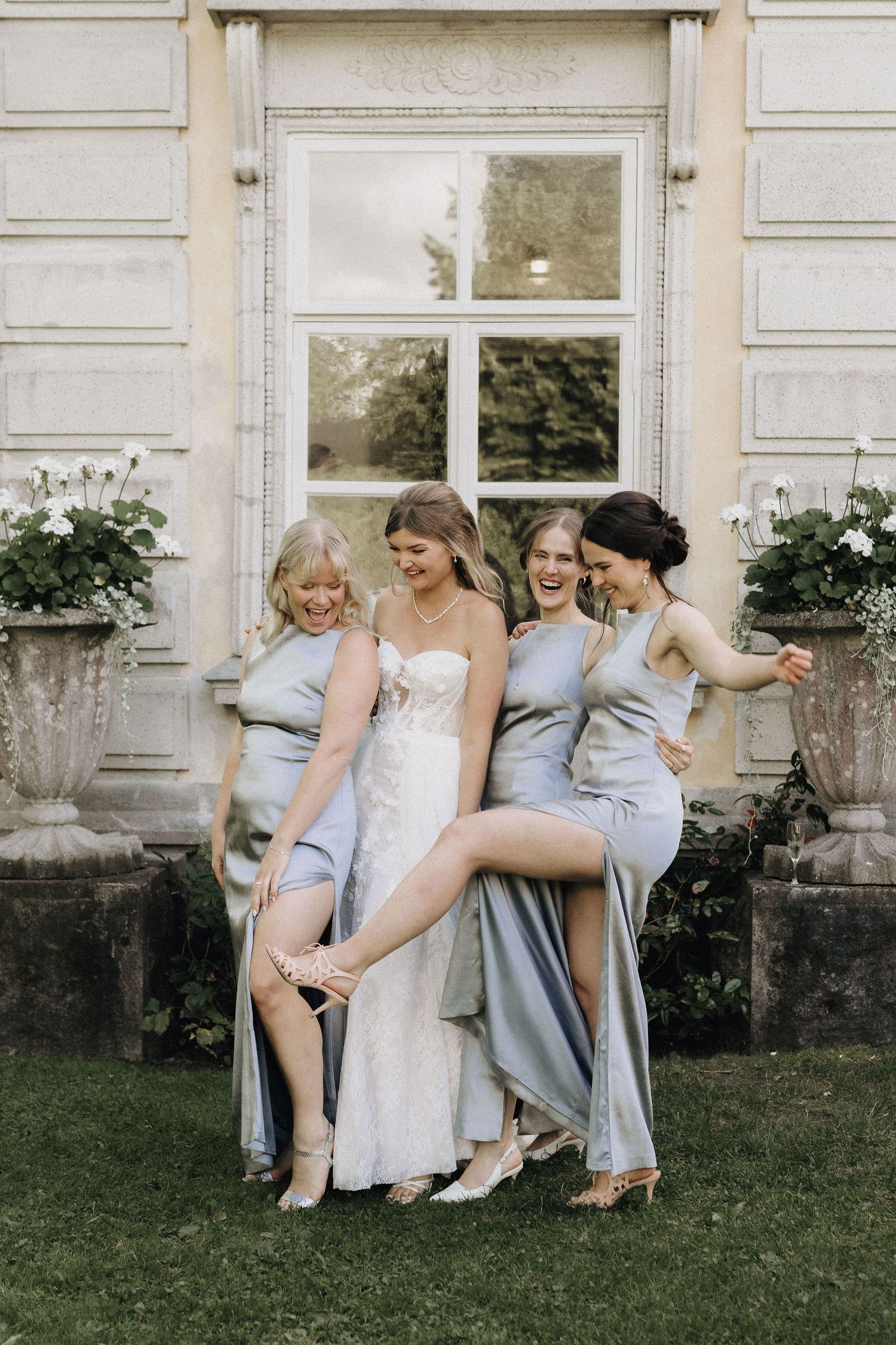 Bride in strapless lace gown laughing with three bridesmaids in blue satin dresses outdoors.