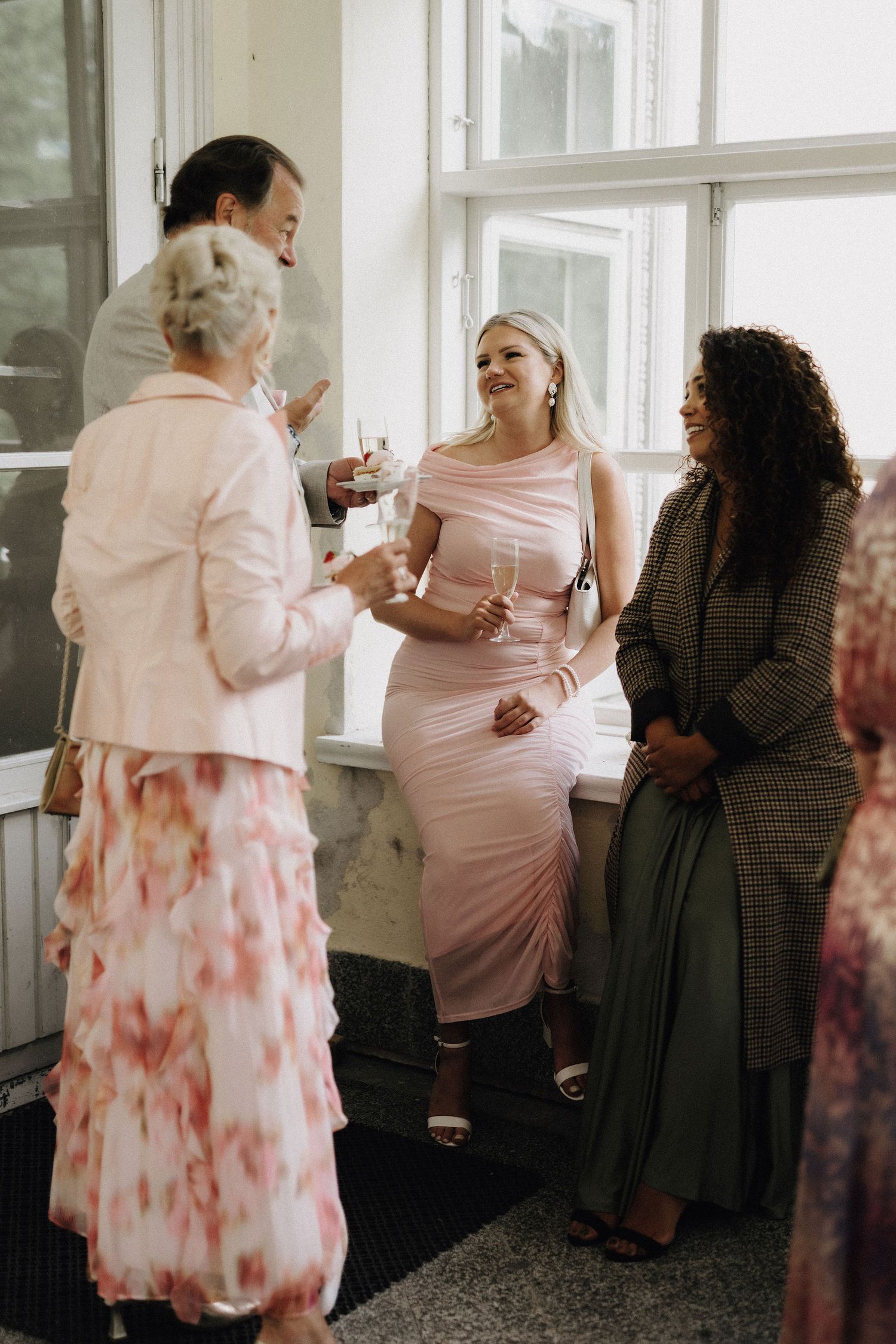 Wedding guests in elegant attire chatting and holding champagne glasses near bright windows at reception.
