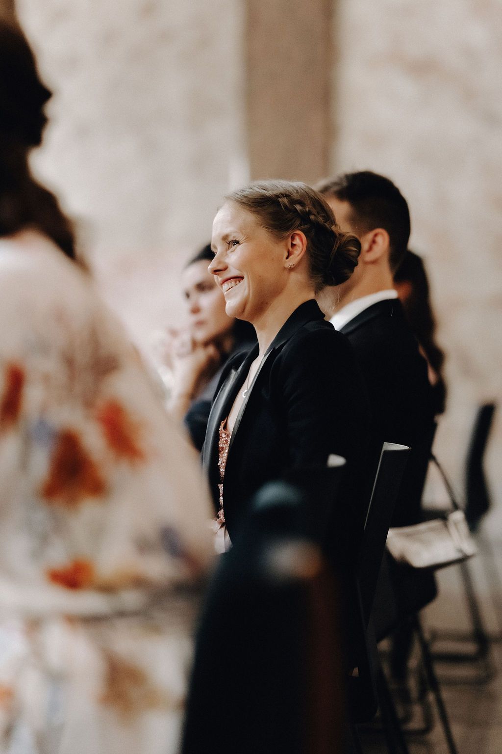 Smiling female wedding guest in black attire enjoys the ceremony among other seated guests.