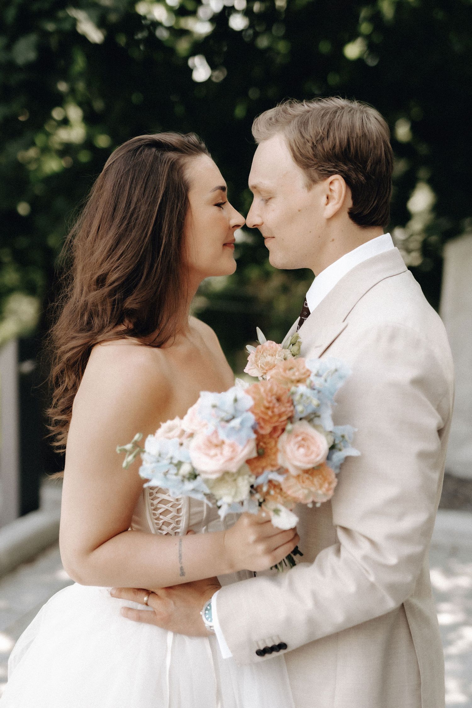 Bride and groom nearly kissing outdoors, she holds a blue and peach bouquet while wearing a strapless white gown.