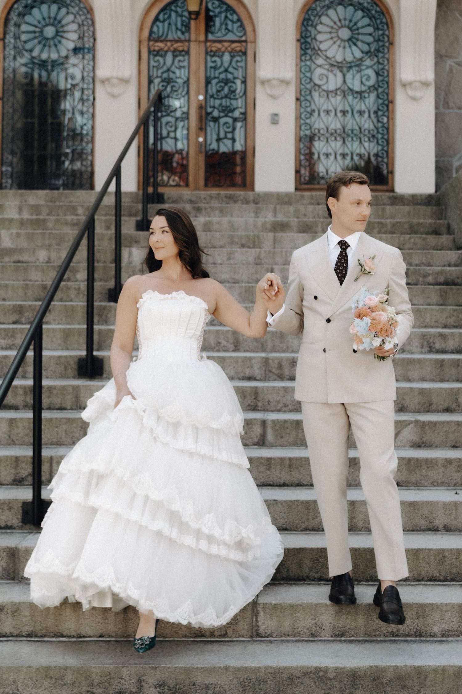 Bride in layered white gown and groom in beige suit descending steps of an ornate building together.
