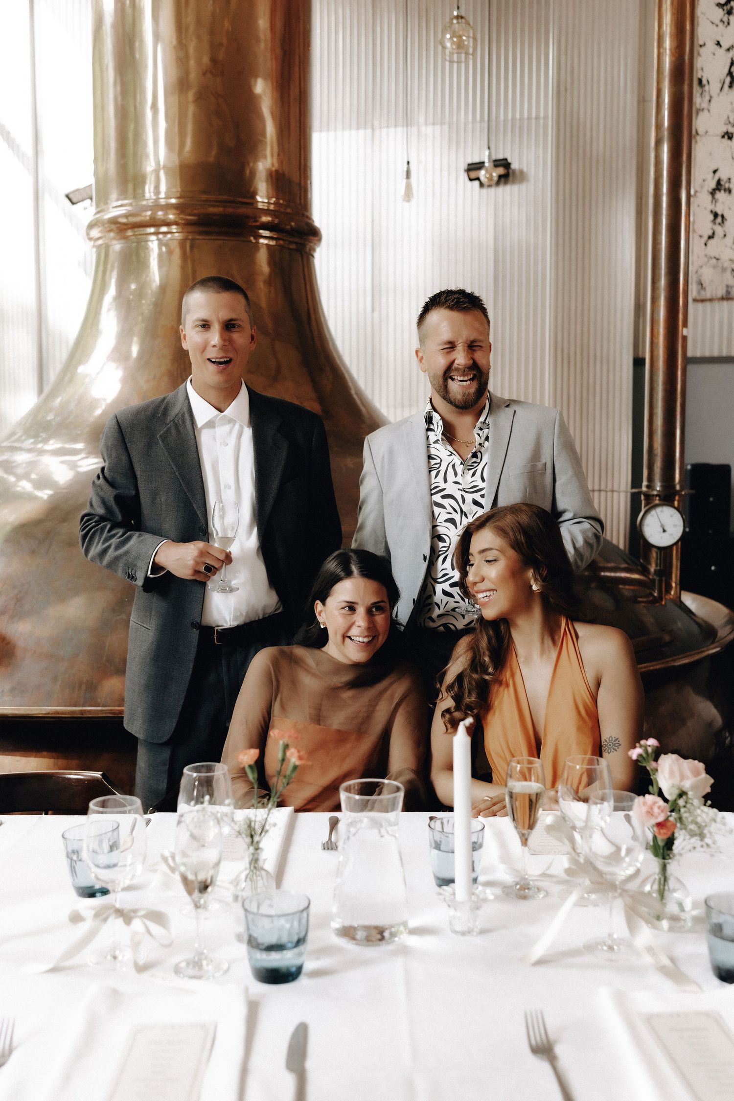 Four wedding guests laughing and celebrating at an elegantly set dinner table in a brewery venue.