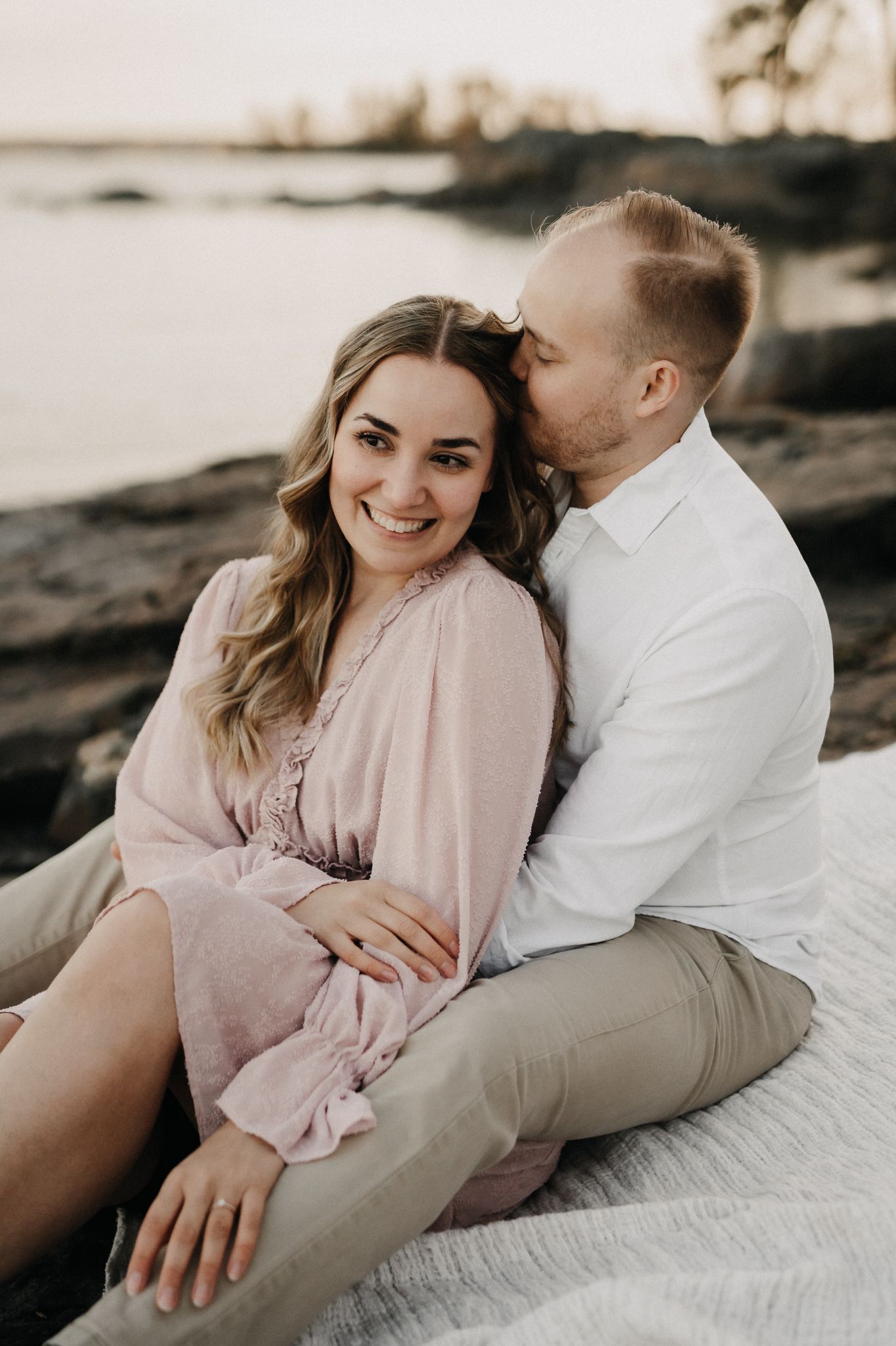 Man kissing woman on the head while she smiles warmly, sitting together on rocks by the water.