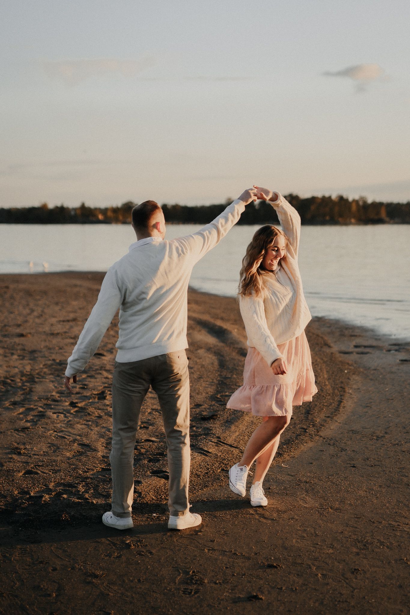 Couple dances romantically on a sandy beach at sunset, man spinning woman in a pink dress by the water.