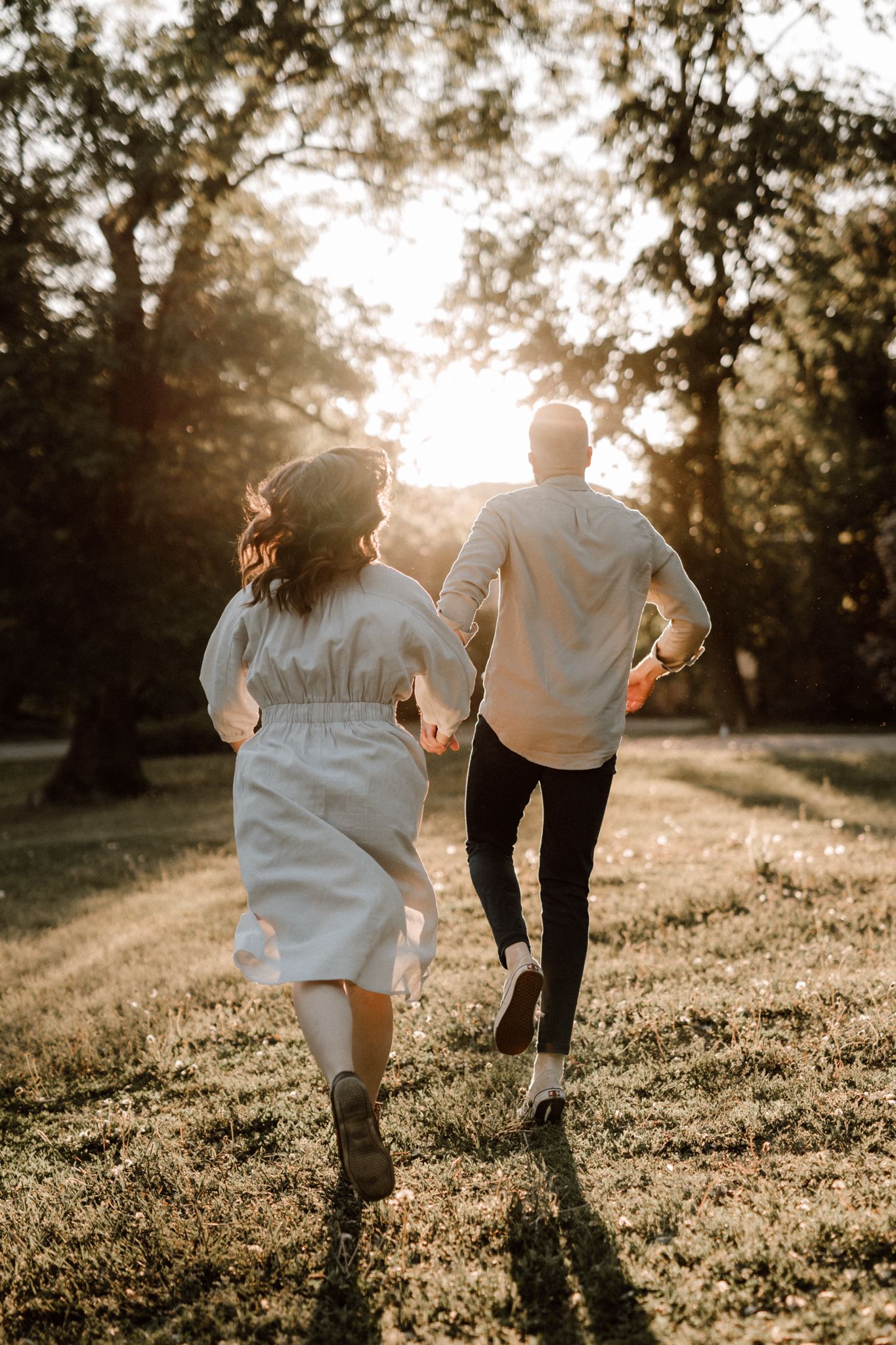 Couple holding hands running away through a sunlit park at golden hour, rear view.