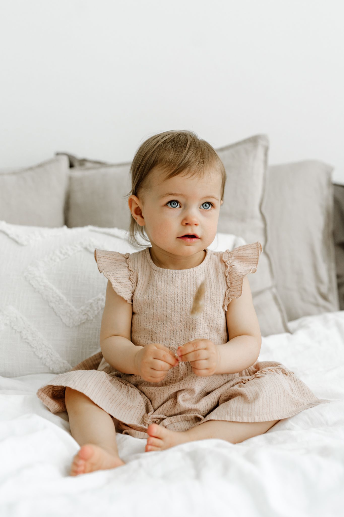 Blue-eyed baby girl in beige ruffled dress sitting on white bed with gray pillows.