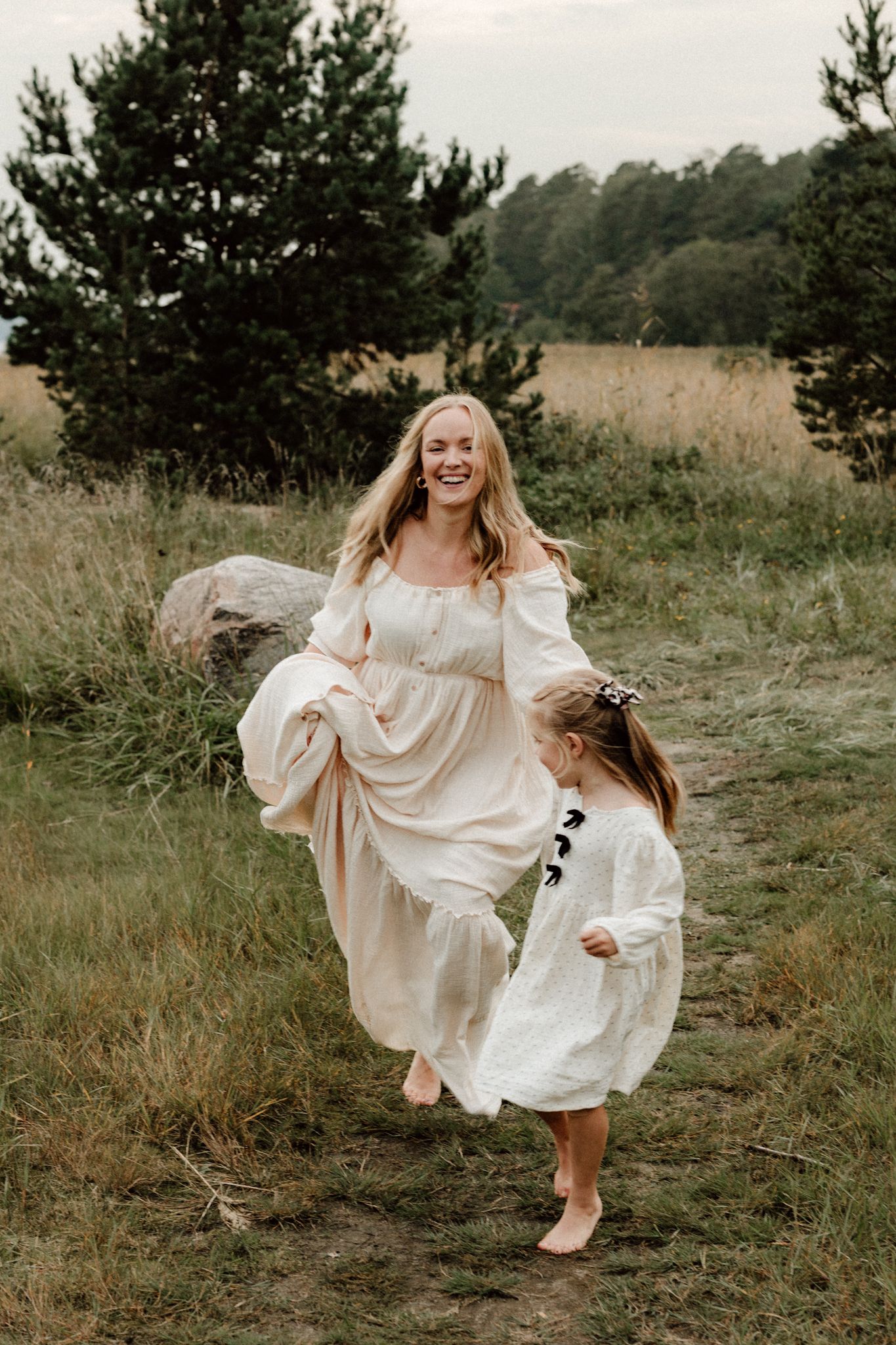 A laughing blonde mother in a cream dress runs playfully with her young daughter in a scenic field.