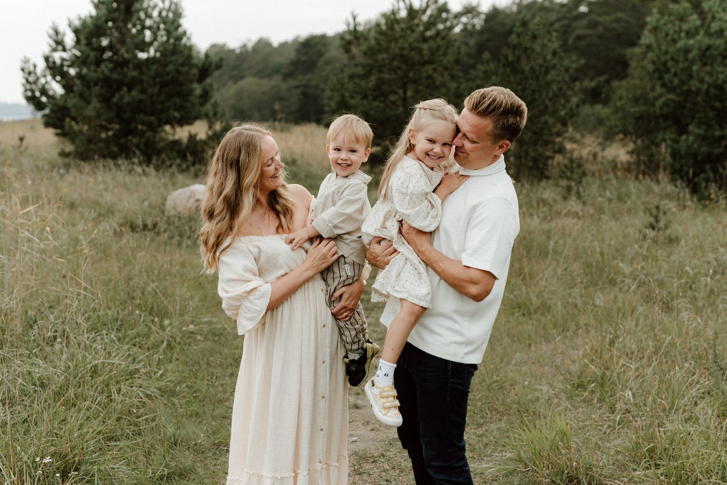 Happy family of four with two toddlers posing in grassy field on cloudy day.