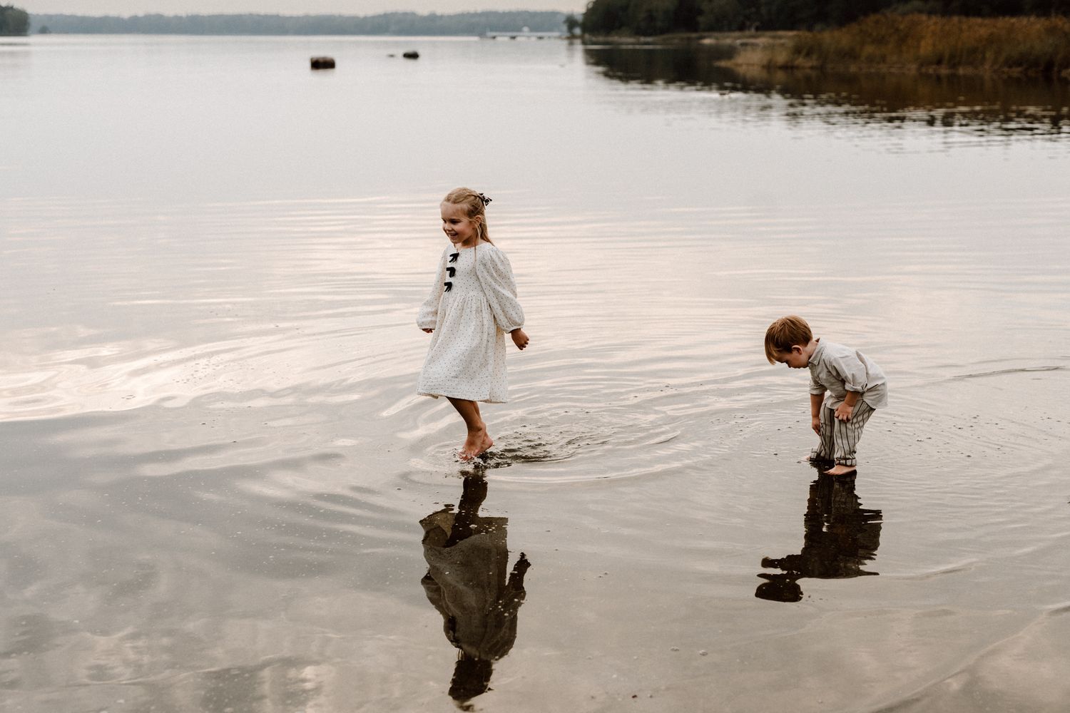 Two young children in white outfits wade and play in the shallow calm waters of a serene lake.