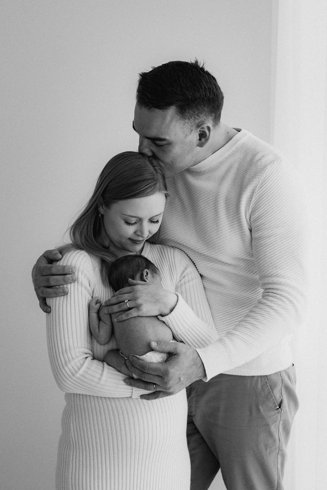 Black and white photo of parents embracing while mother holds newborn baby.