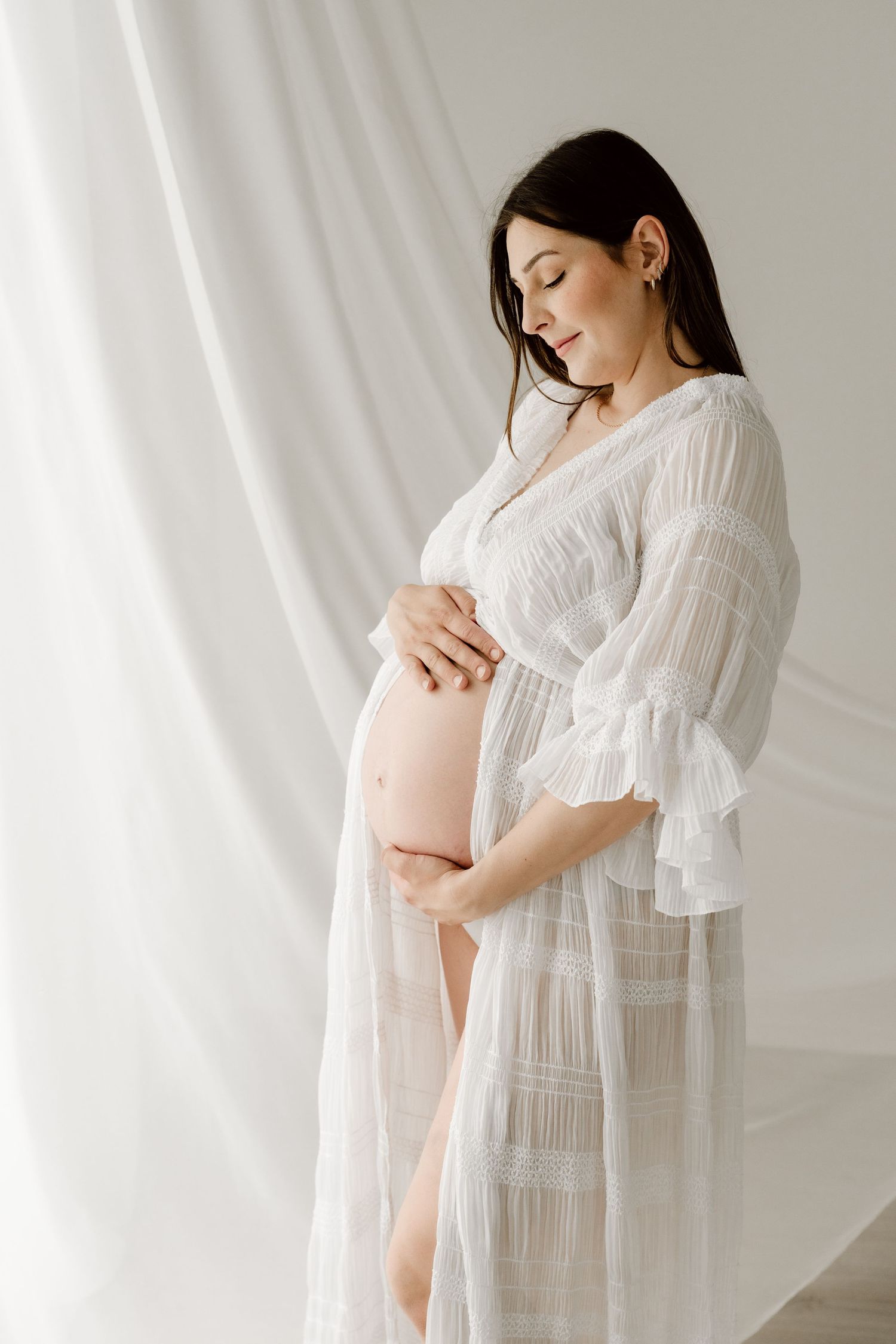 A pregnant woman in a sheer white robe gently cradles her baby bump near flowing white curtains.