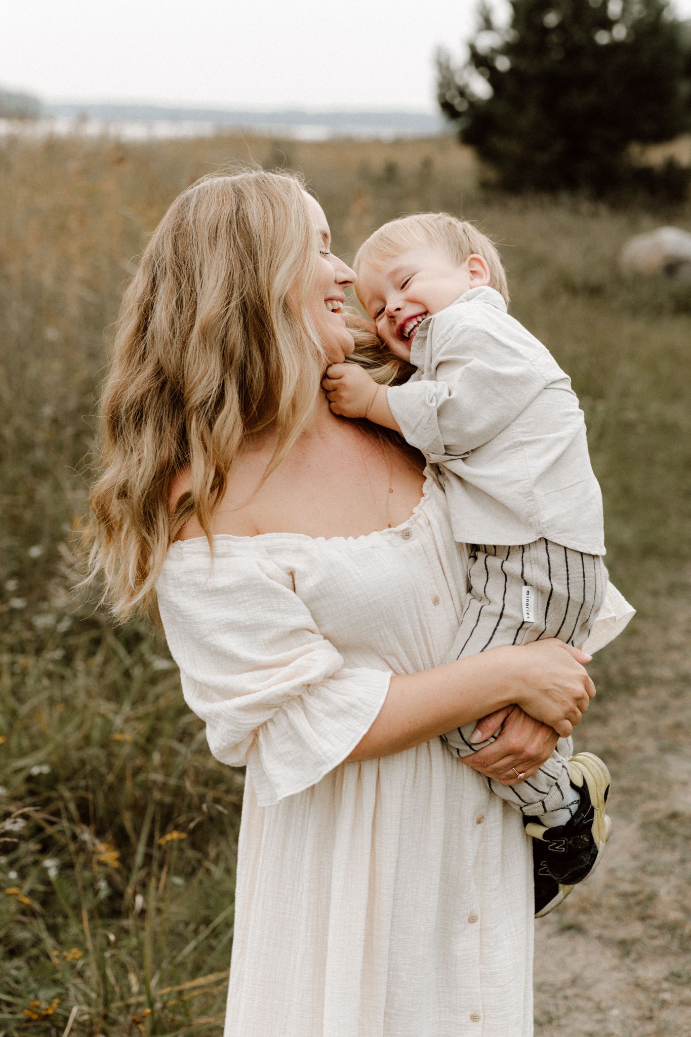 Blonde woman in white off-shoulder dress laughing with joyful toddler boy outdoors.