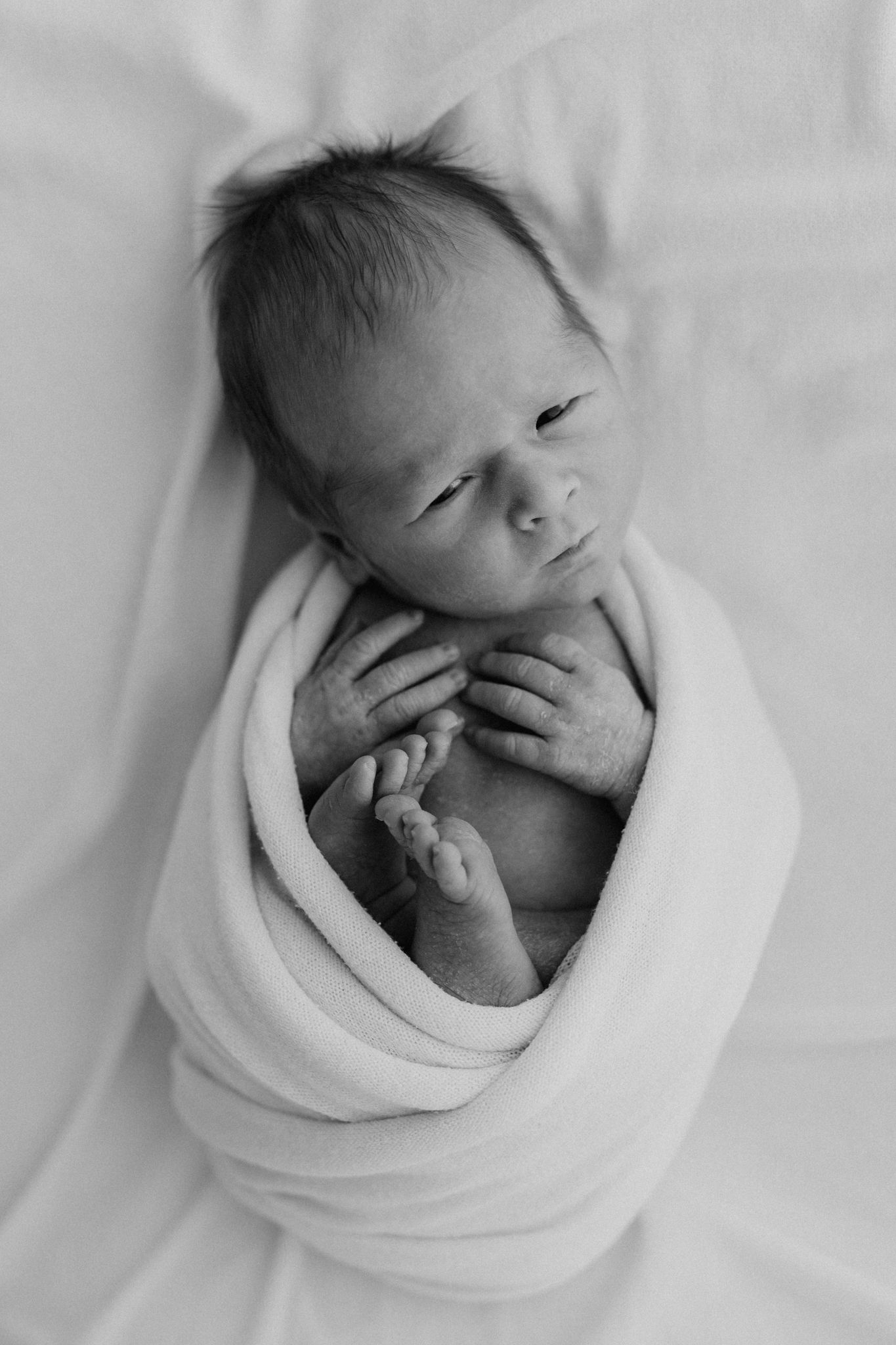Black and white overhead view of a sleeping newborn baby swaddled snugly in a soft white blanket.