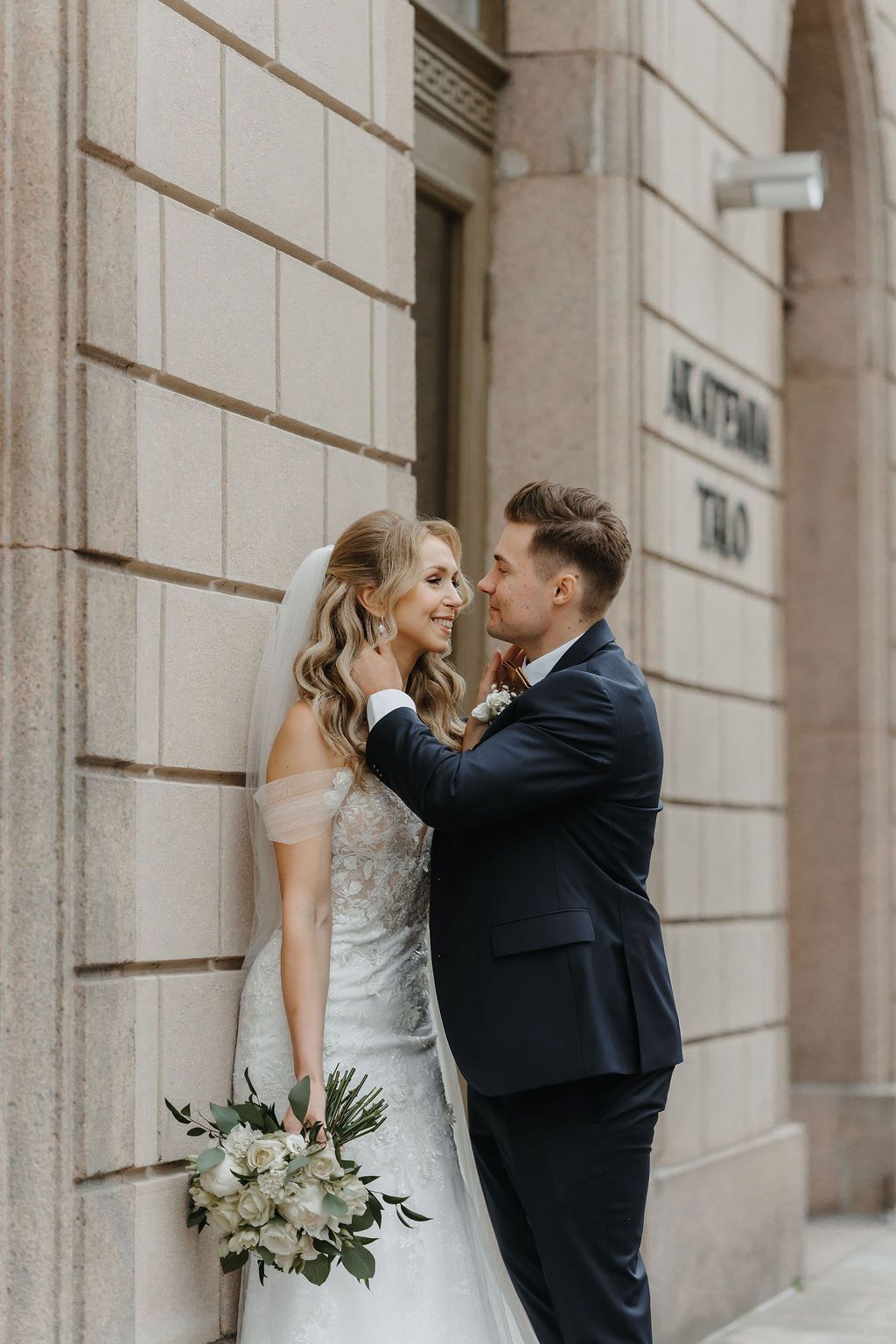 Groom gently touching bride's face as they share a romantic moment against Akatemia-Talo stone wall.