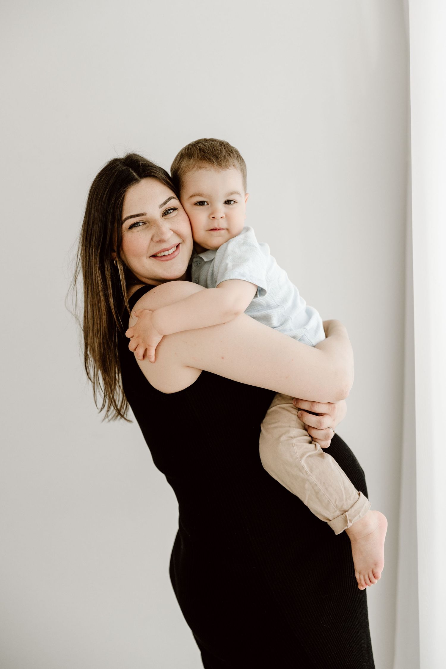 A smiling brunette mother in a black dress lovingly holds her toddler son against a white background.