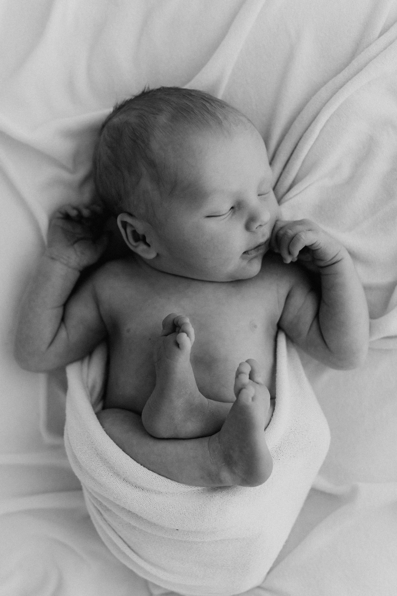 Black and white photo of a sleeping newborn baby loosely swaddled in a white blanket on soft fabric.