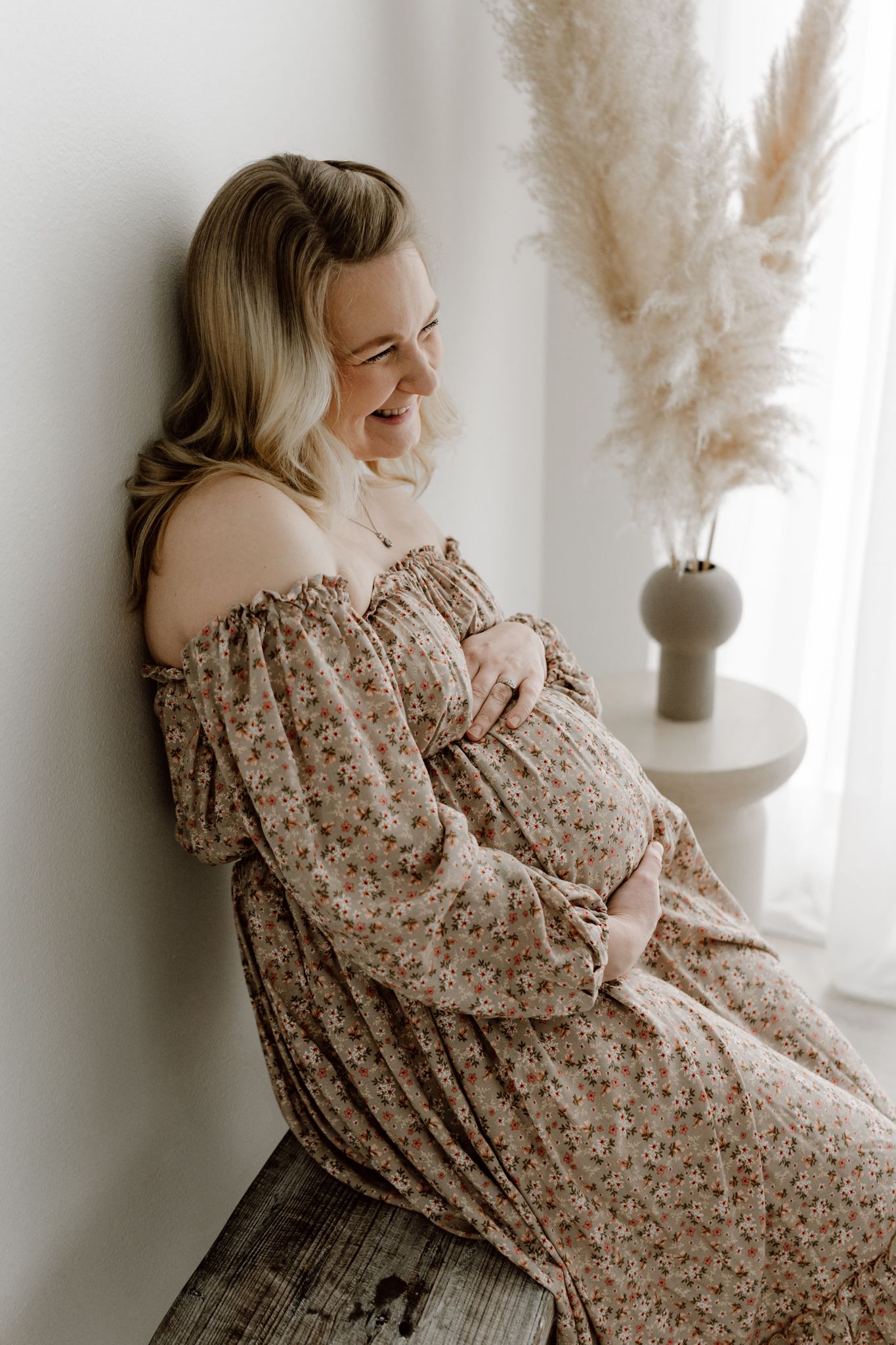 Glowing pregnant woman in floral off-shoulder dress laughing while seated, pampas grass vase beside her in studio.