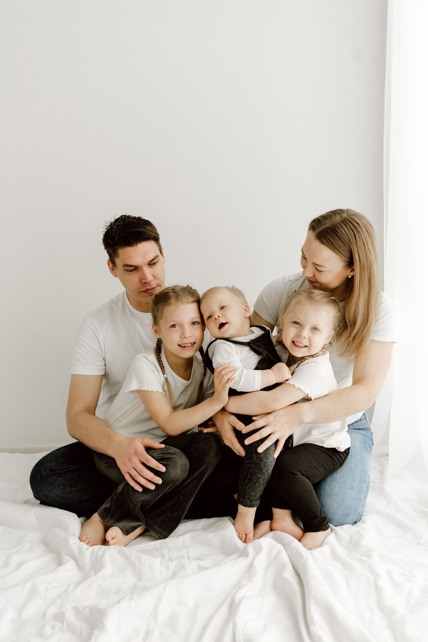 Happy family of five sitting together on white bed, parents with three young children.