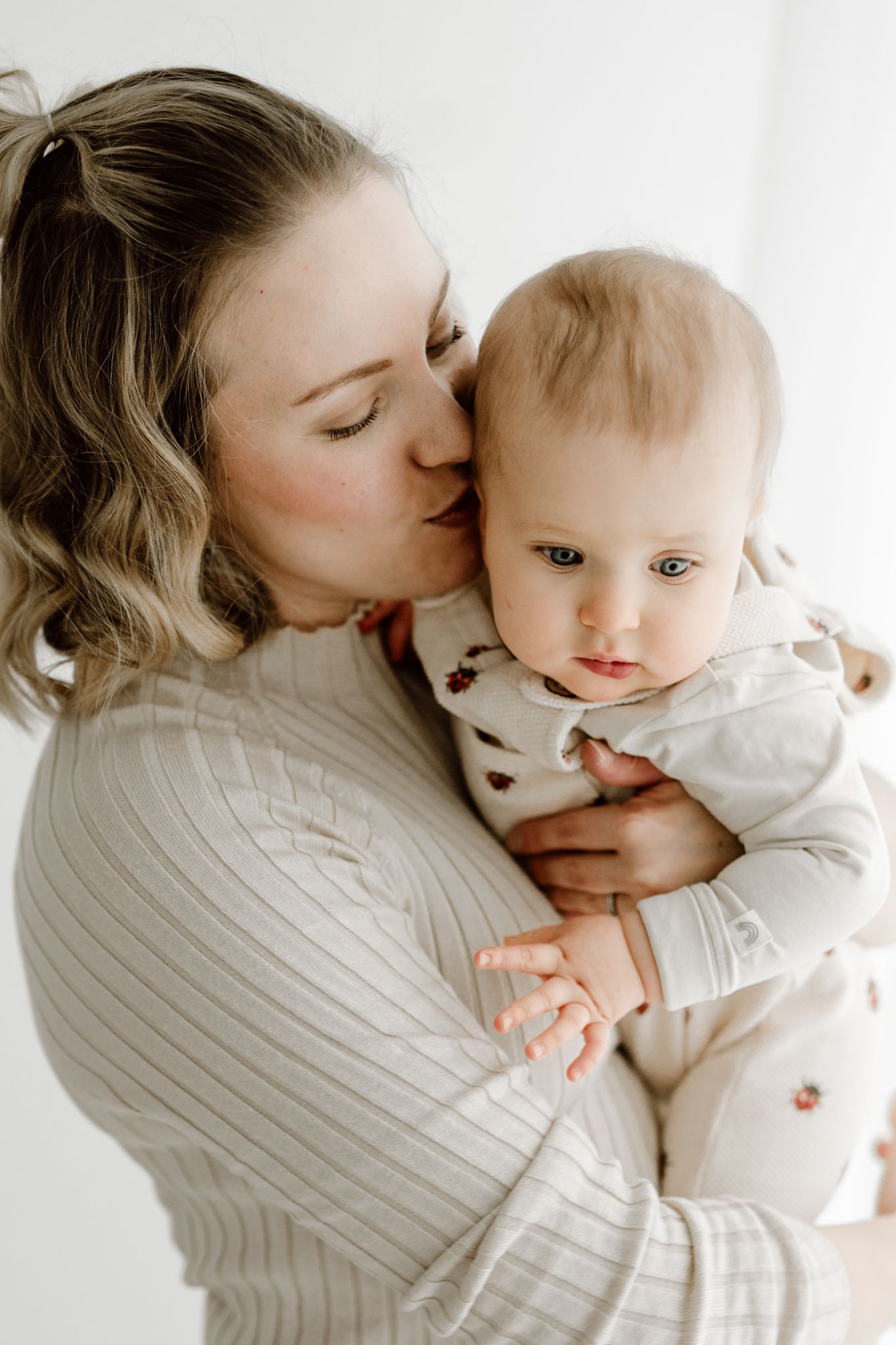 Tender moment of a mother kissing her calm baby on the cheek indoors.