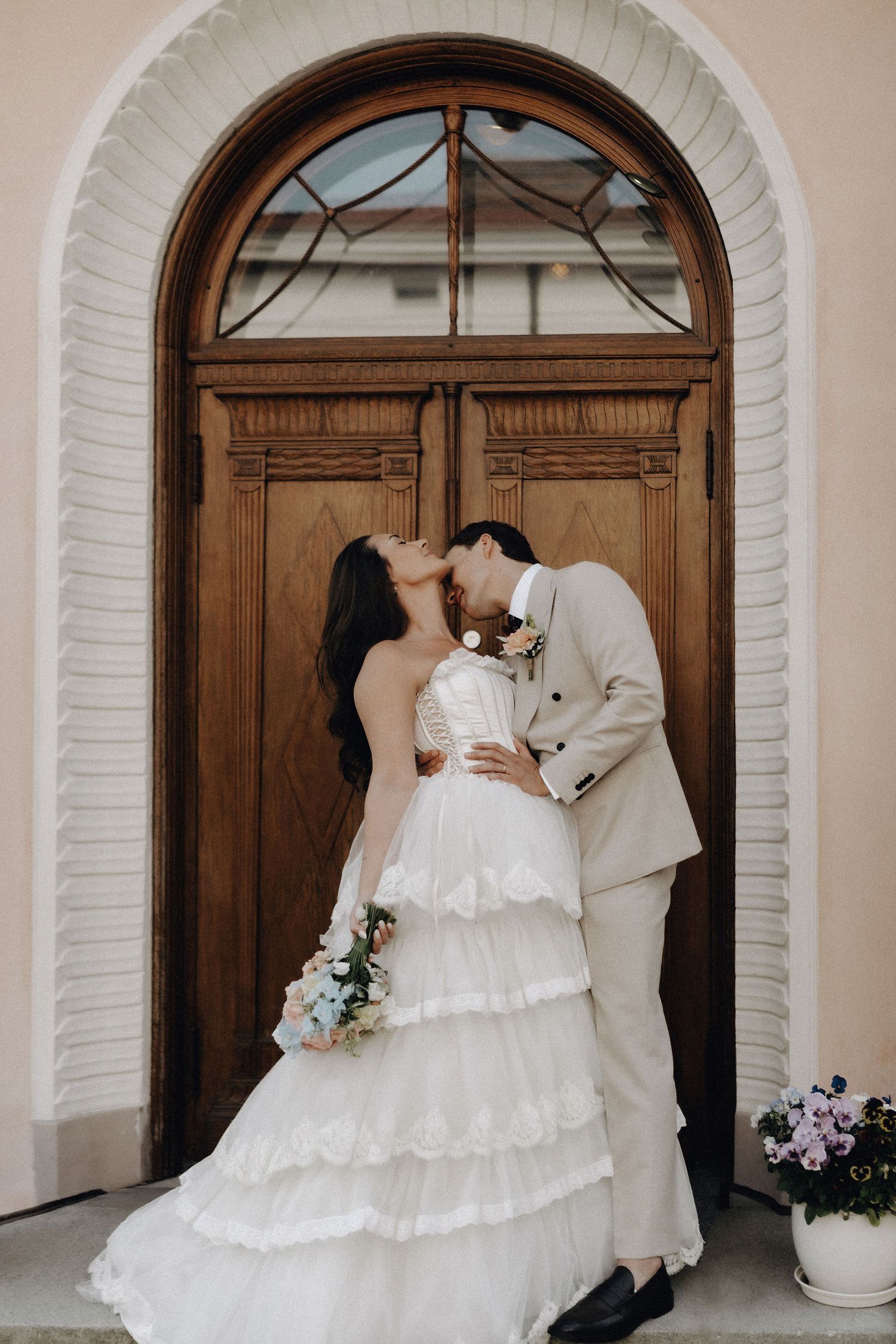 Couple shares a passionate kiss in front of a classic arched wooden door on their wedding day.