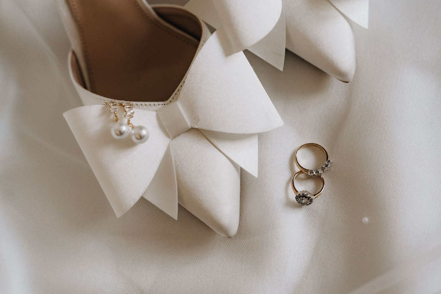 White satin bridal heels with pearl bow detail beside gold wedding rings on soft fabric.