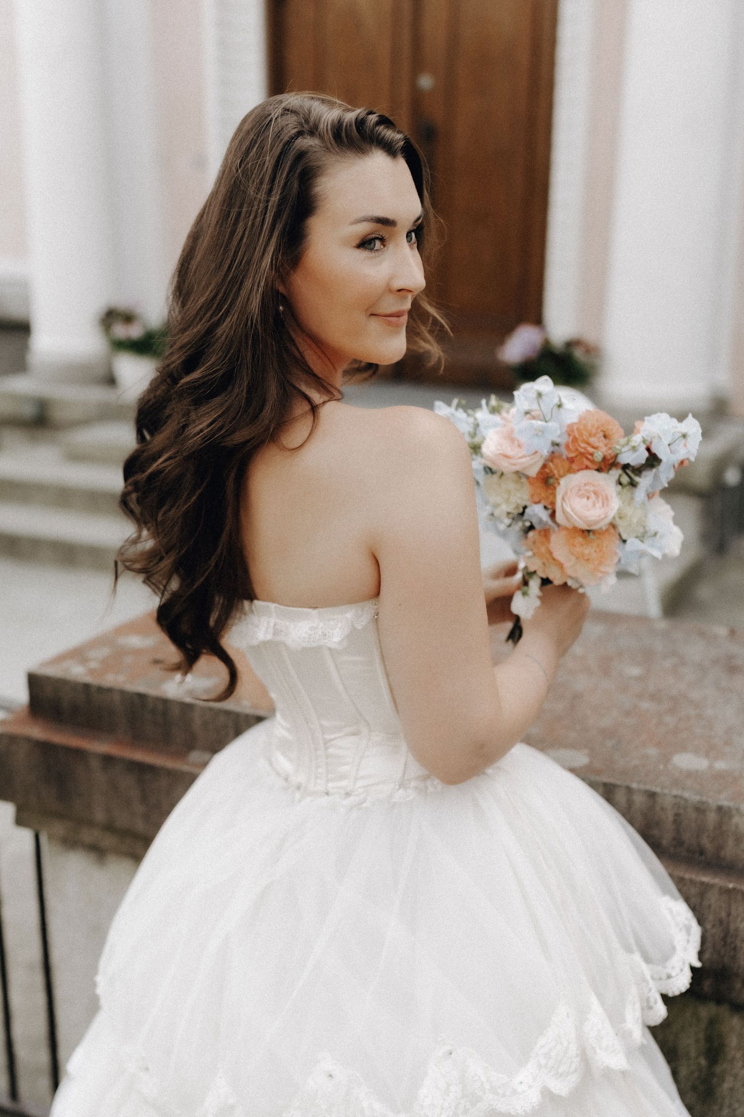 Bride with long brunette hair in a strapless white corset gown holding a peach and blue bouquet, looking back.