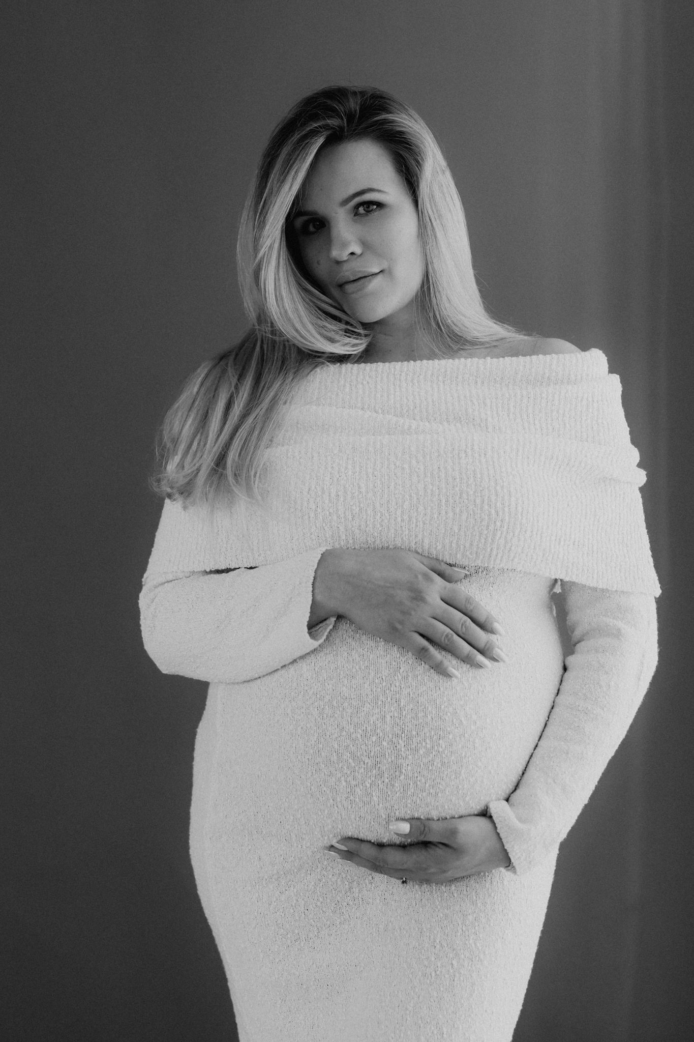 Black and white maternity photo of pregnant blonde woman in white off-shoulder dress holding baby bump.