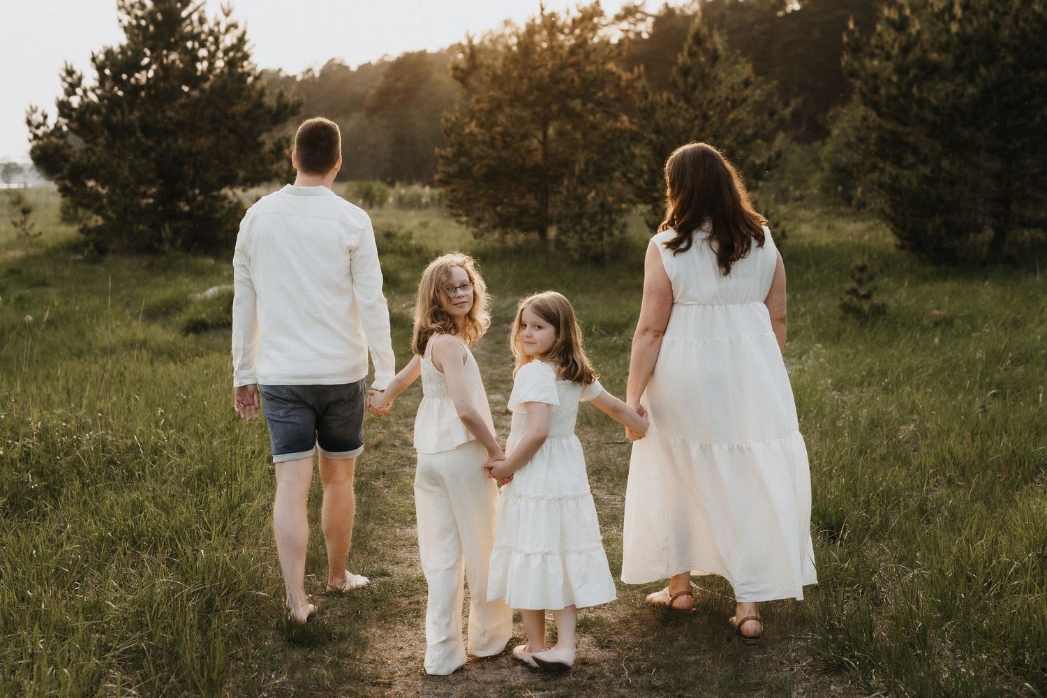 A family of four walks hand in hand through a sunny green meadow wearing coordinated white outfits.