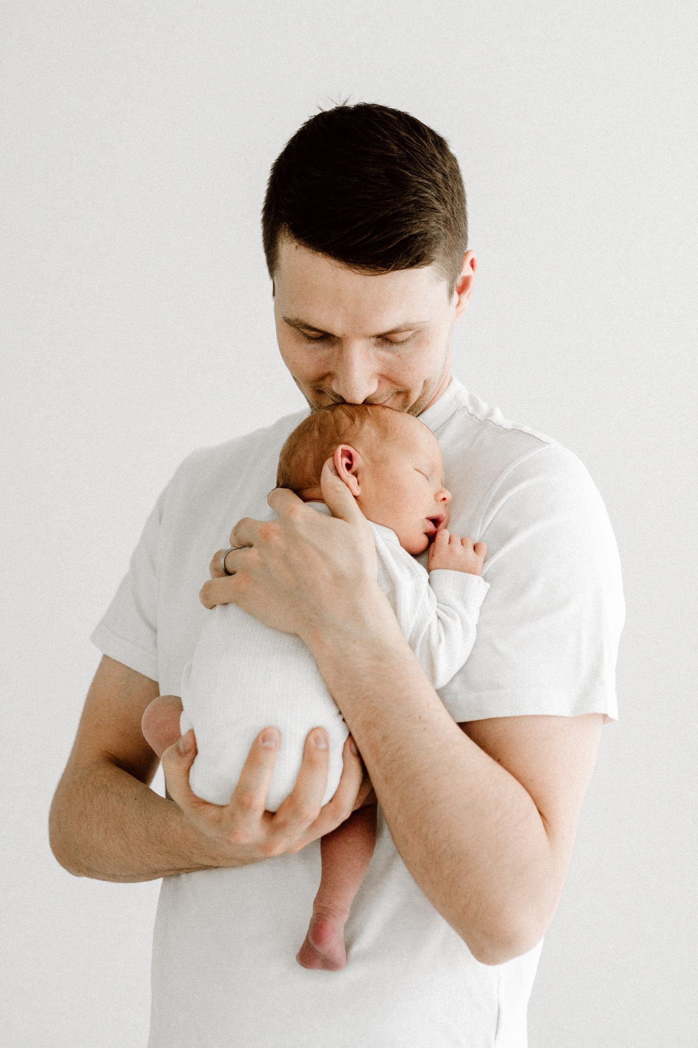 Father in white t-shirt gently kissing his sleeping newborn baby held against his chest.