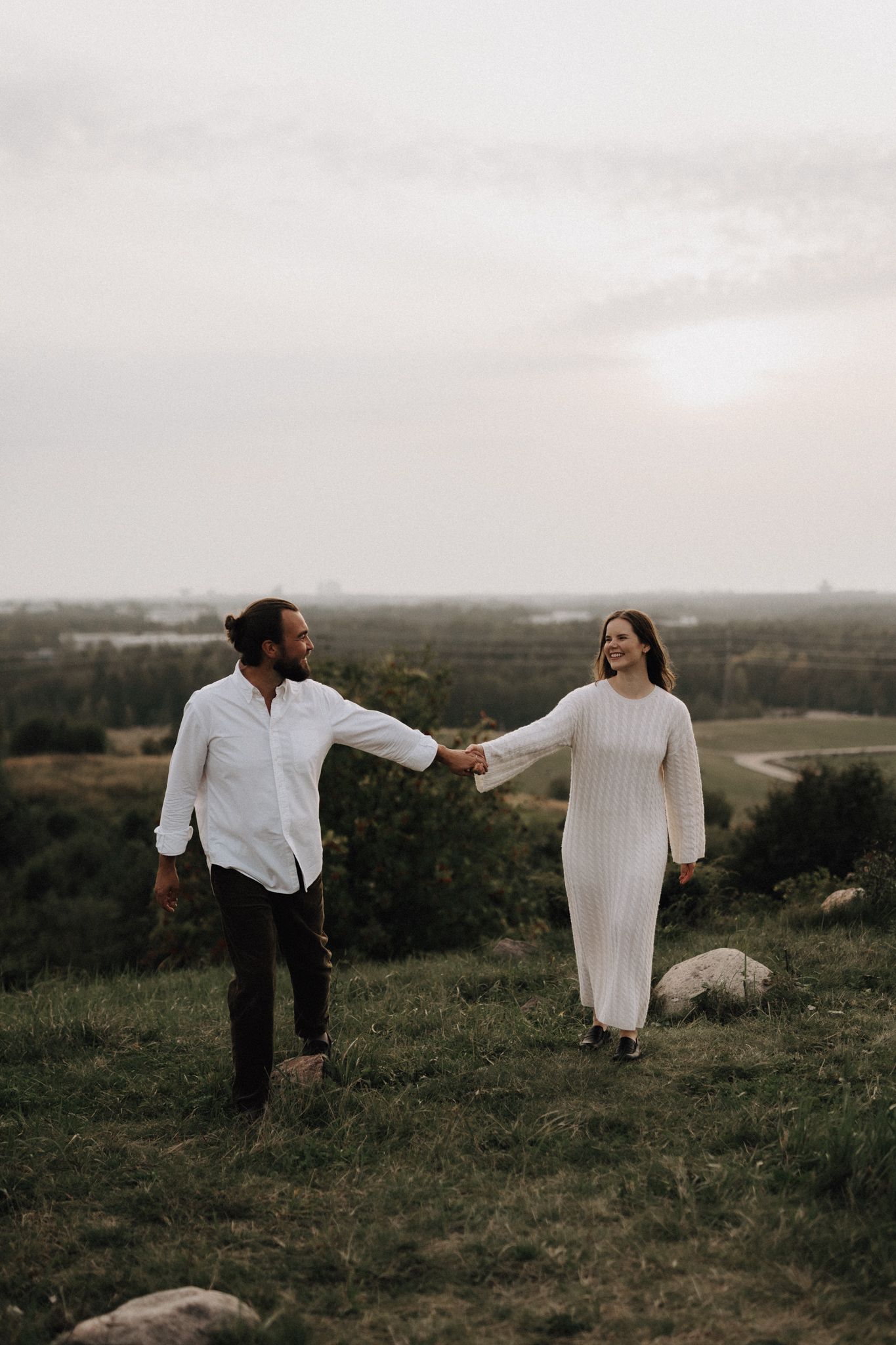 A laughing couple in white outfits holds hands on a grassy hill with a vast landscape and cloudy sky behind.