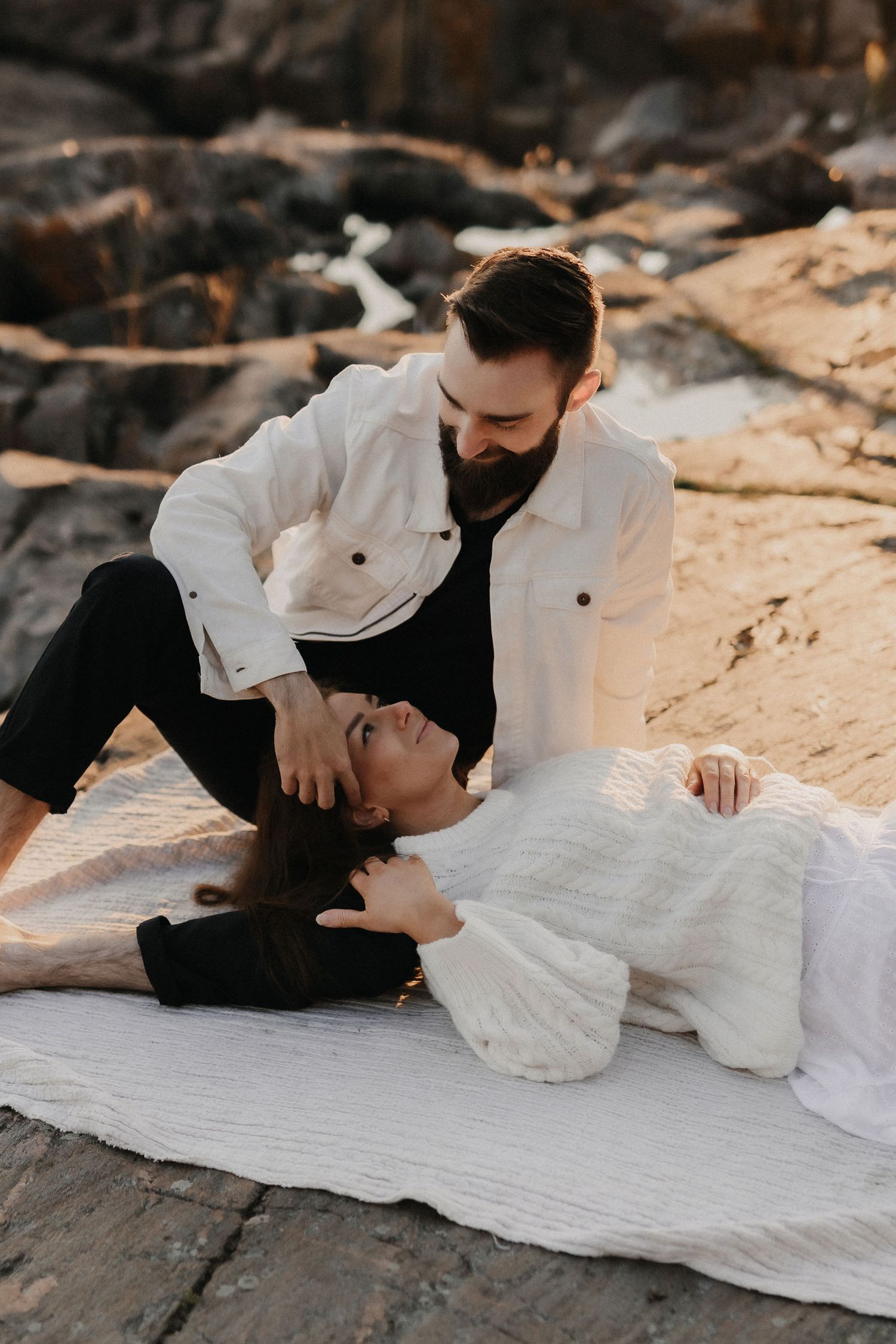 Bearded man tenderly gazing at woman lying on white blanket on rocky beach at sunset.
