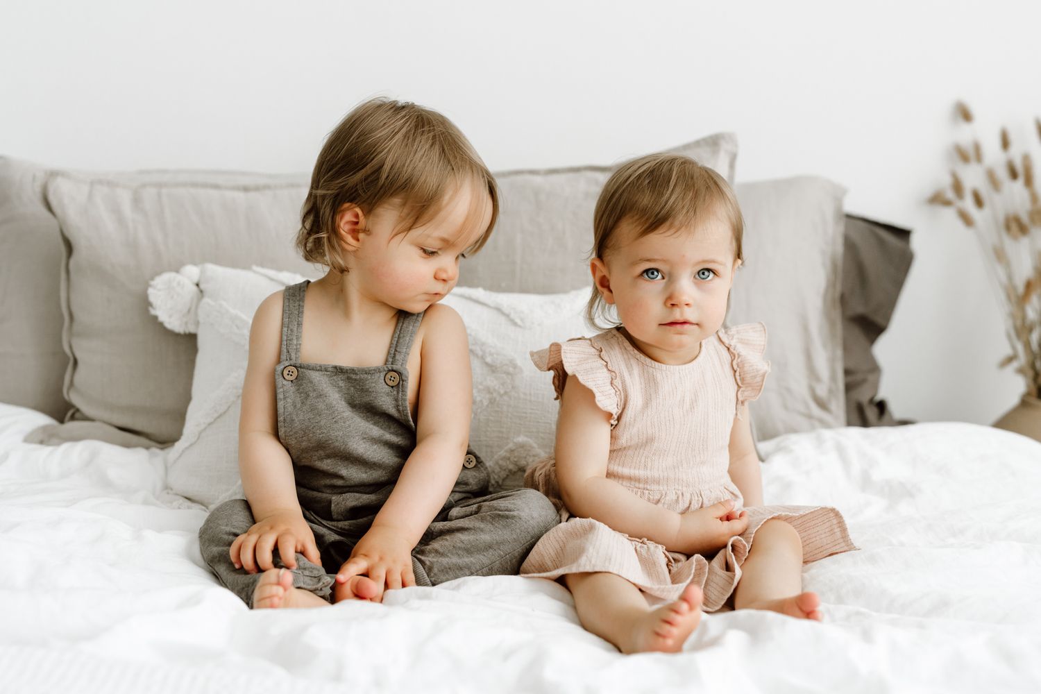 Two baby girls in neutral outfits sitting together on white bedding with gray pillows.