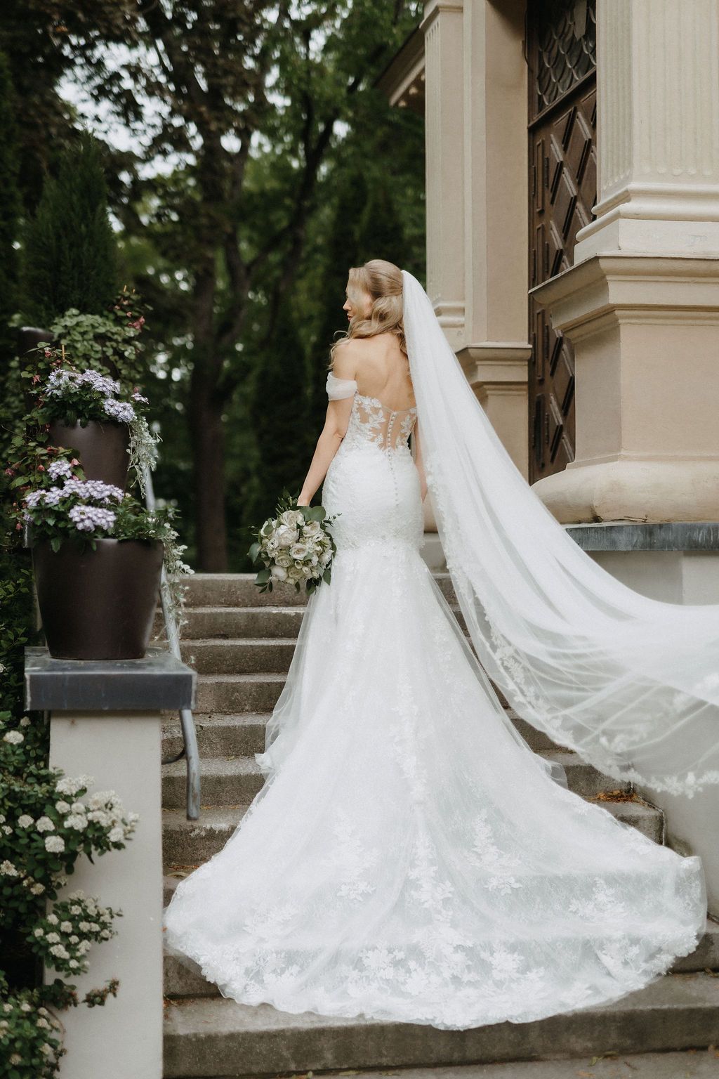 Bride in backless lace mermaid gown with flowing cathedral veil descends stone steps of classical venue.