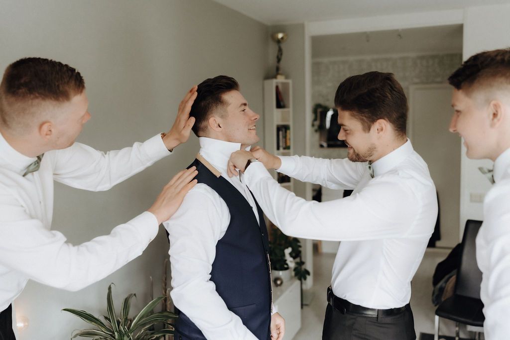 Groomsmen in white shirts helping groom adjust his tie and collar while laughing.