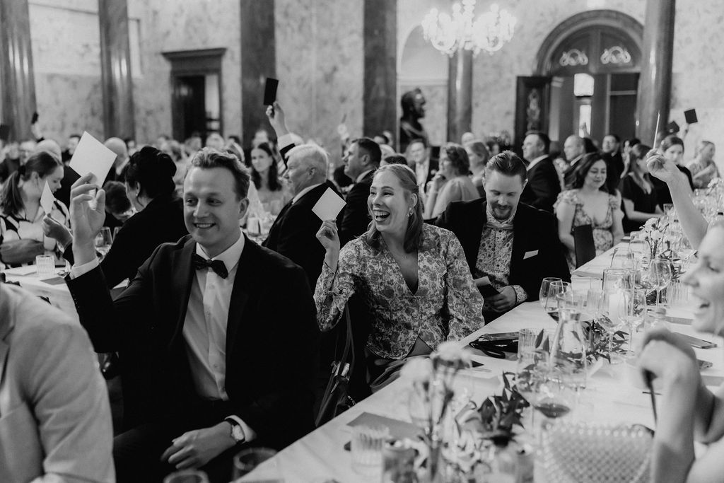 Wedding guests laugh and celebrate during speeches at a formal black-tie reception.