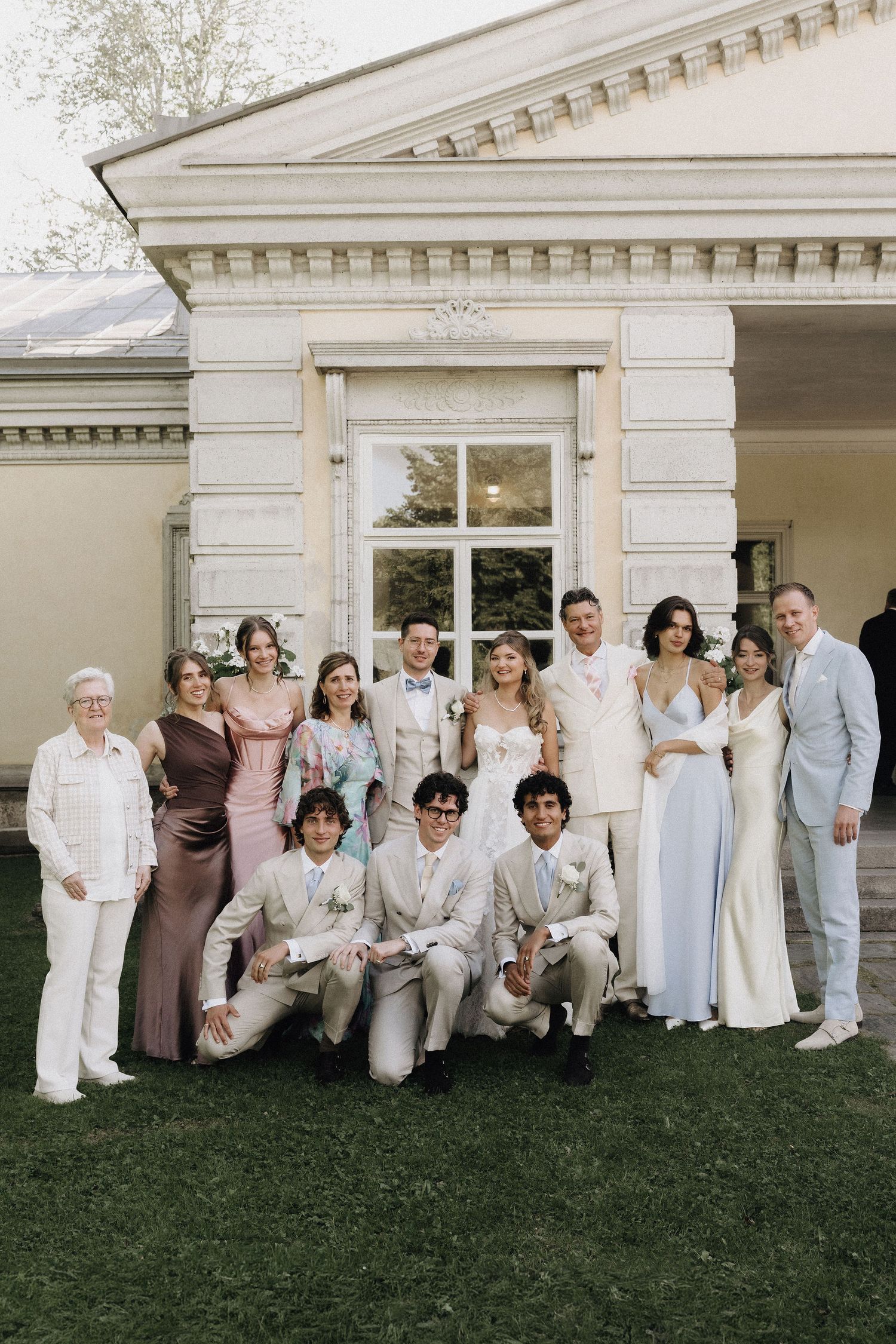 Large wedding party group photo outside elegant venue with bride in white gown and guests in formal attire.